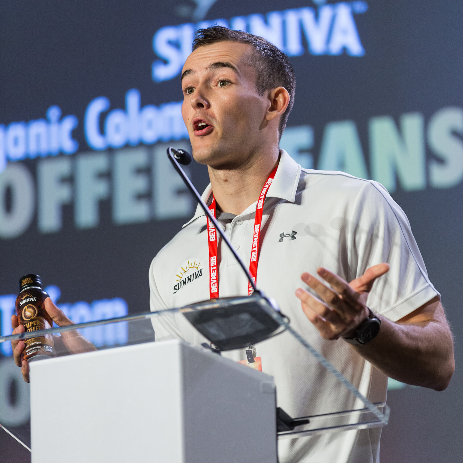 A man in a white Sunniva polo holds a bottle of Sunniva Super Coffee below the lectern while a screen behind him reads Sunniva Organic Colombian Coffee Beans.