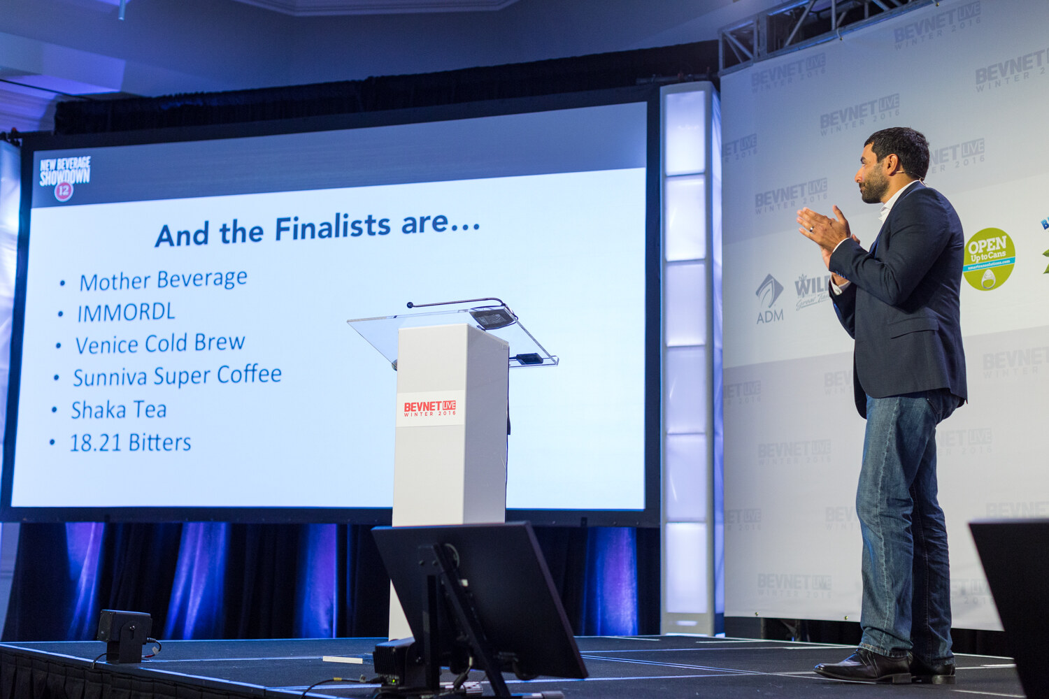 A man in a navy blazer and jeans claps on stage beside a screen reading And the Finalists are, listing Mother Beverage, IMMORDL, Venice Cold Brew, Sunniva Super Coffee, Shaka Tea and 18.21 Bitters.