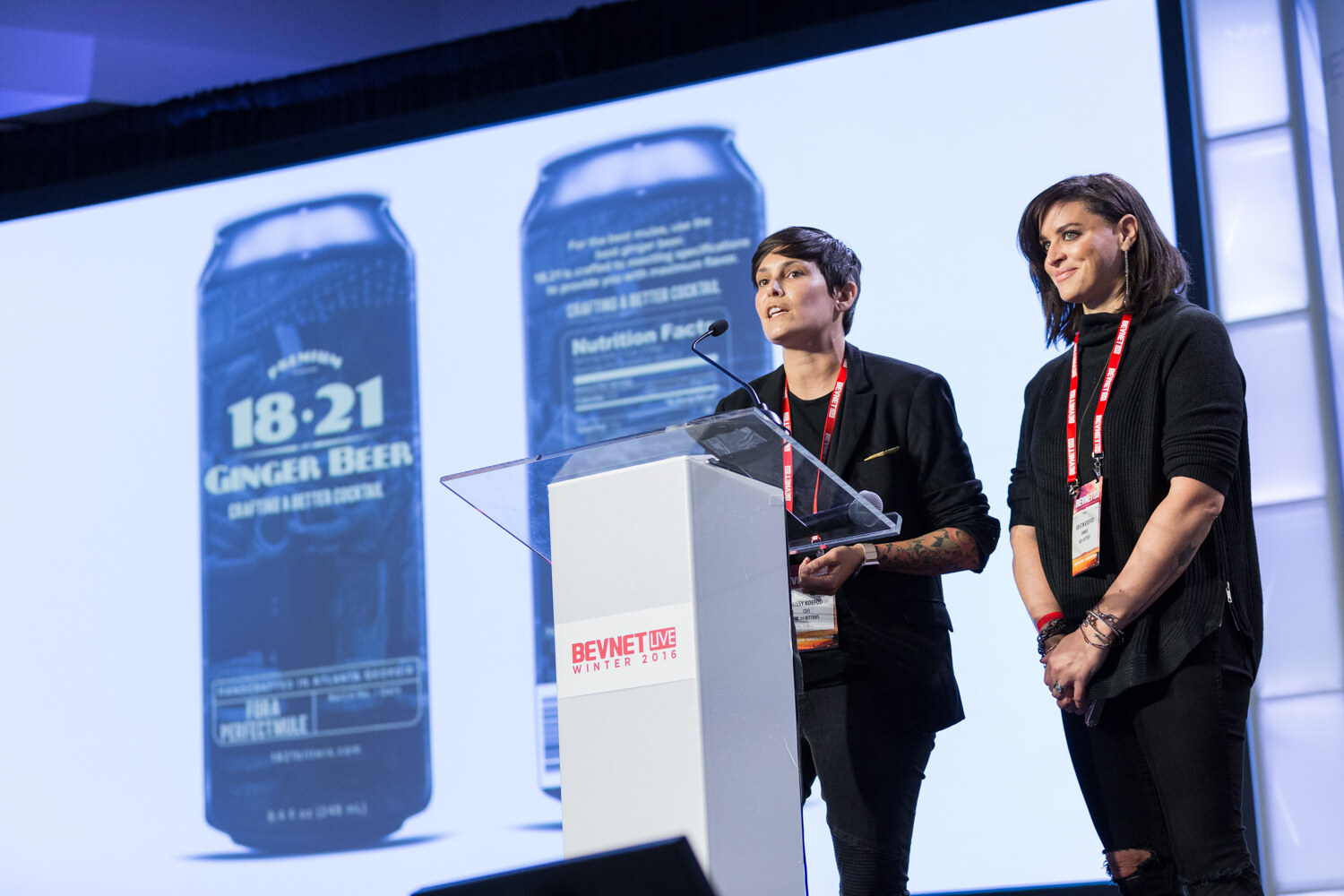 Two people in black stand at a clear lectern, one speaking into the microphone, in front of a large slide showing cans labeled 18.21 Ginger Beer.