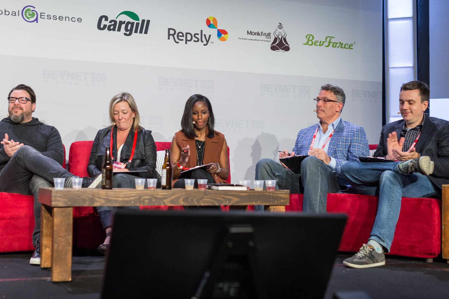 Five people with BevNET Live lanyards sit side by side on red couches behind a wooden table holding sample cups and two brown bottles, under a wall of sponsor logos including Cargill, Repsly and BevForce.