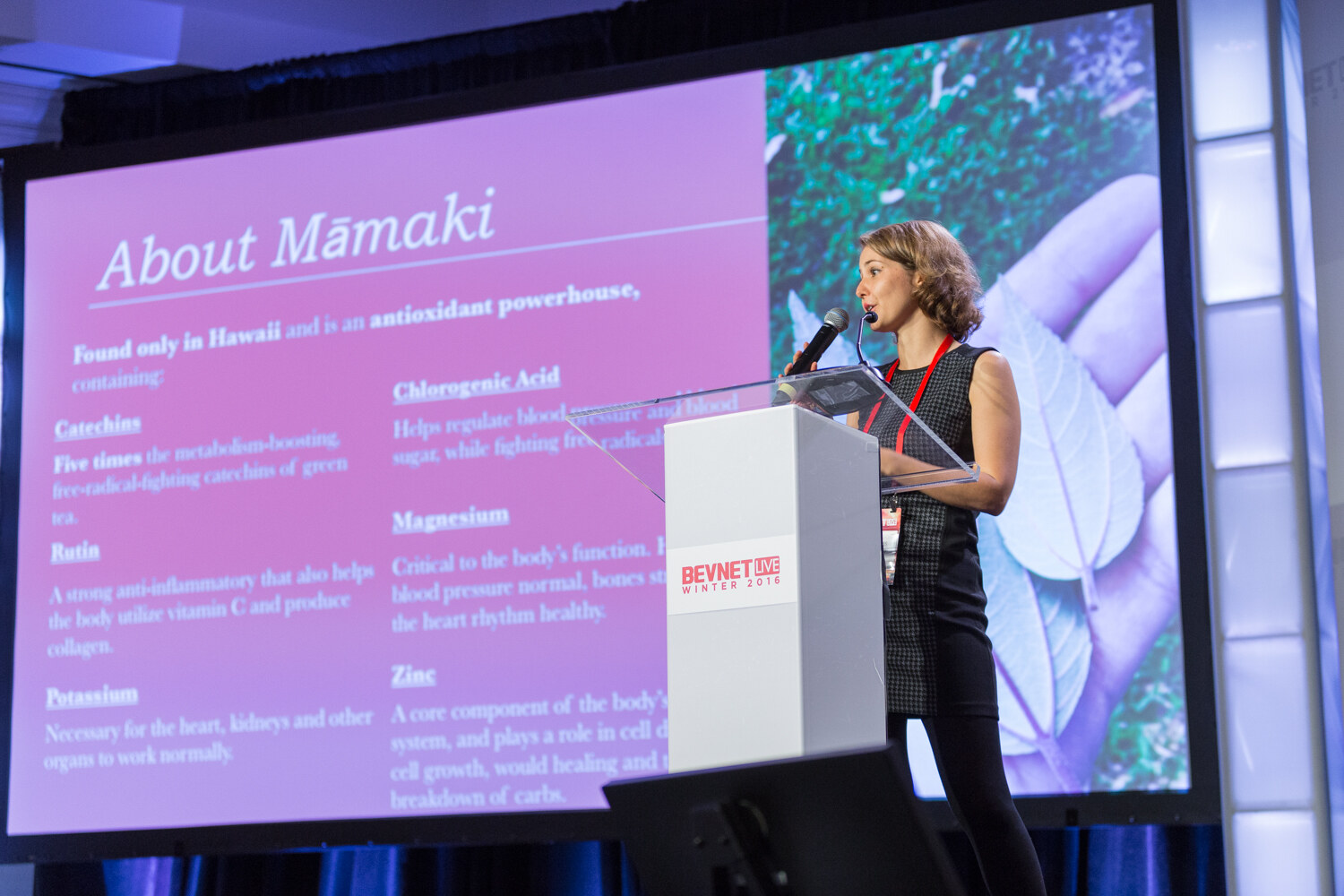 A woman in a gray dress holds a microphone at a clear lectern while the screen shows a pink slide titled About Mamaki, listing catechins, rutin, potassium, chlorogenic acid, magnesium and zinc.