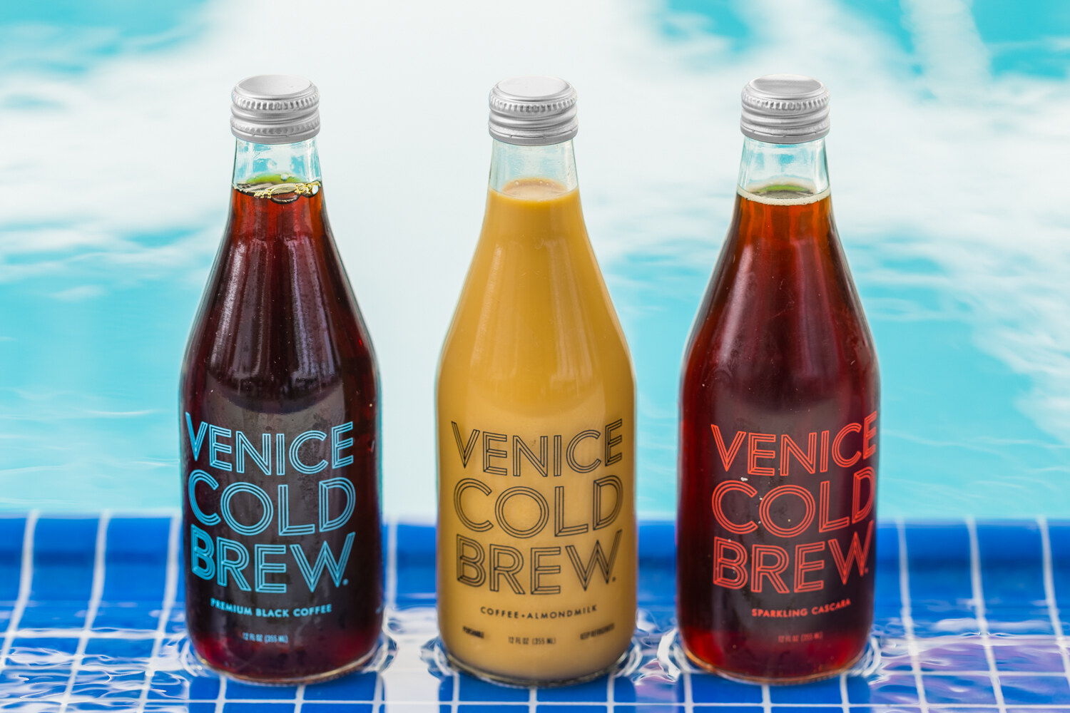 Three glass bottles labeled Venice Cold Brew, in black coffee, coffee-almondmilk and sparkling cascara varieties, stand on a blue-tiled ledge with the pool water behind them.