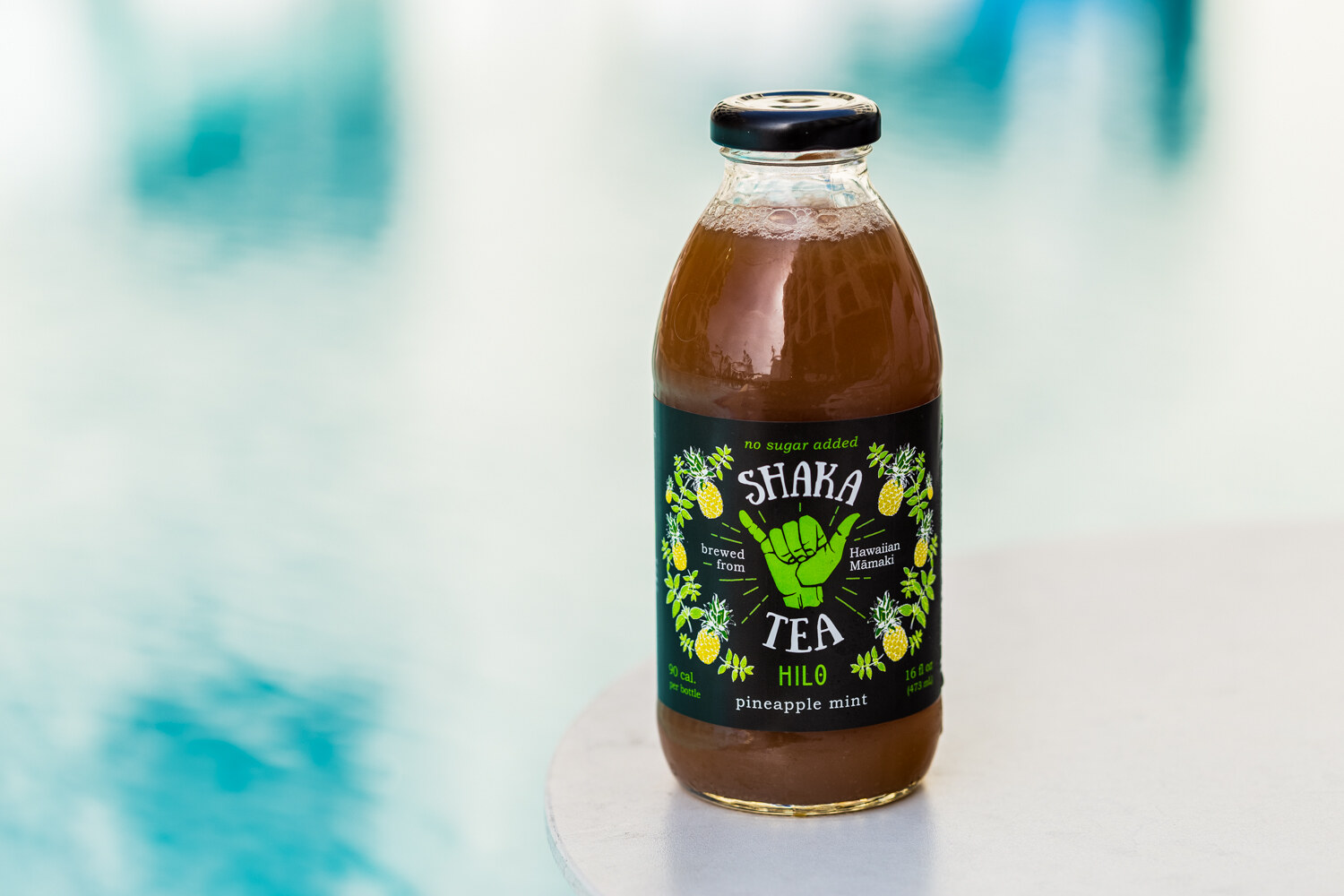 A glass bottle of Shaka Tea Hilo pineapple mint with a green and black label showing a shaka hand sign stands on a white table with a turquoise blur behind it.