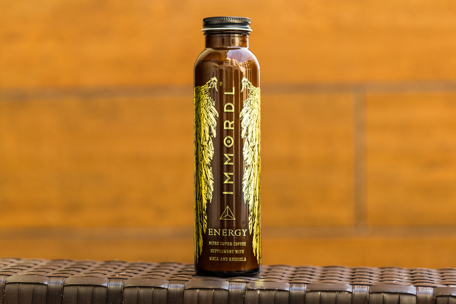 A dark glass bottle with gold winged artwork and the word IMMORDL Energy stands on a woven surface against an orange wooden wall.