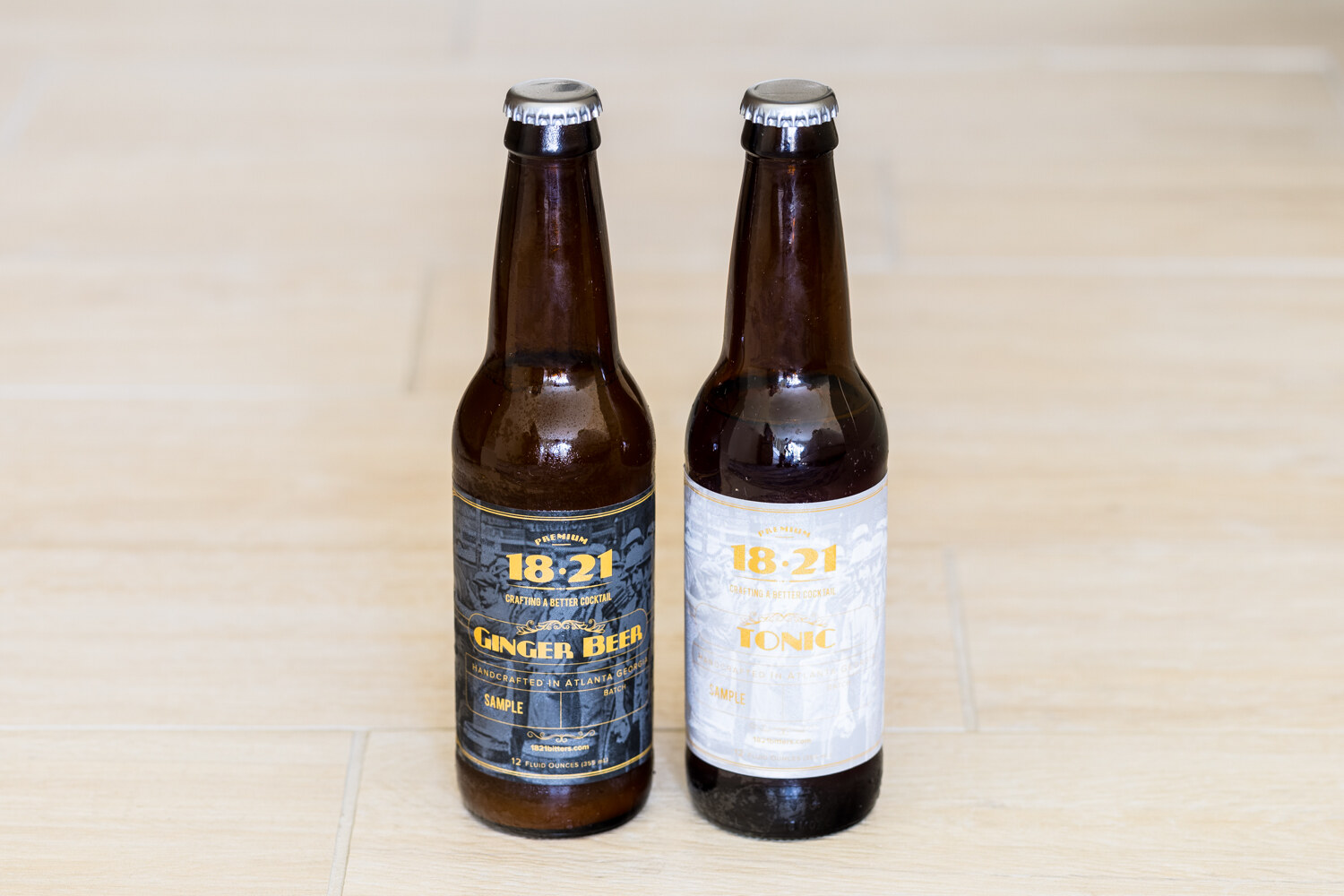 A brown bottle labeled 18.21 Ginger Beer and a brown bottle with a white 18.21 Tonic label stand next to each other on a pale wood floor.