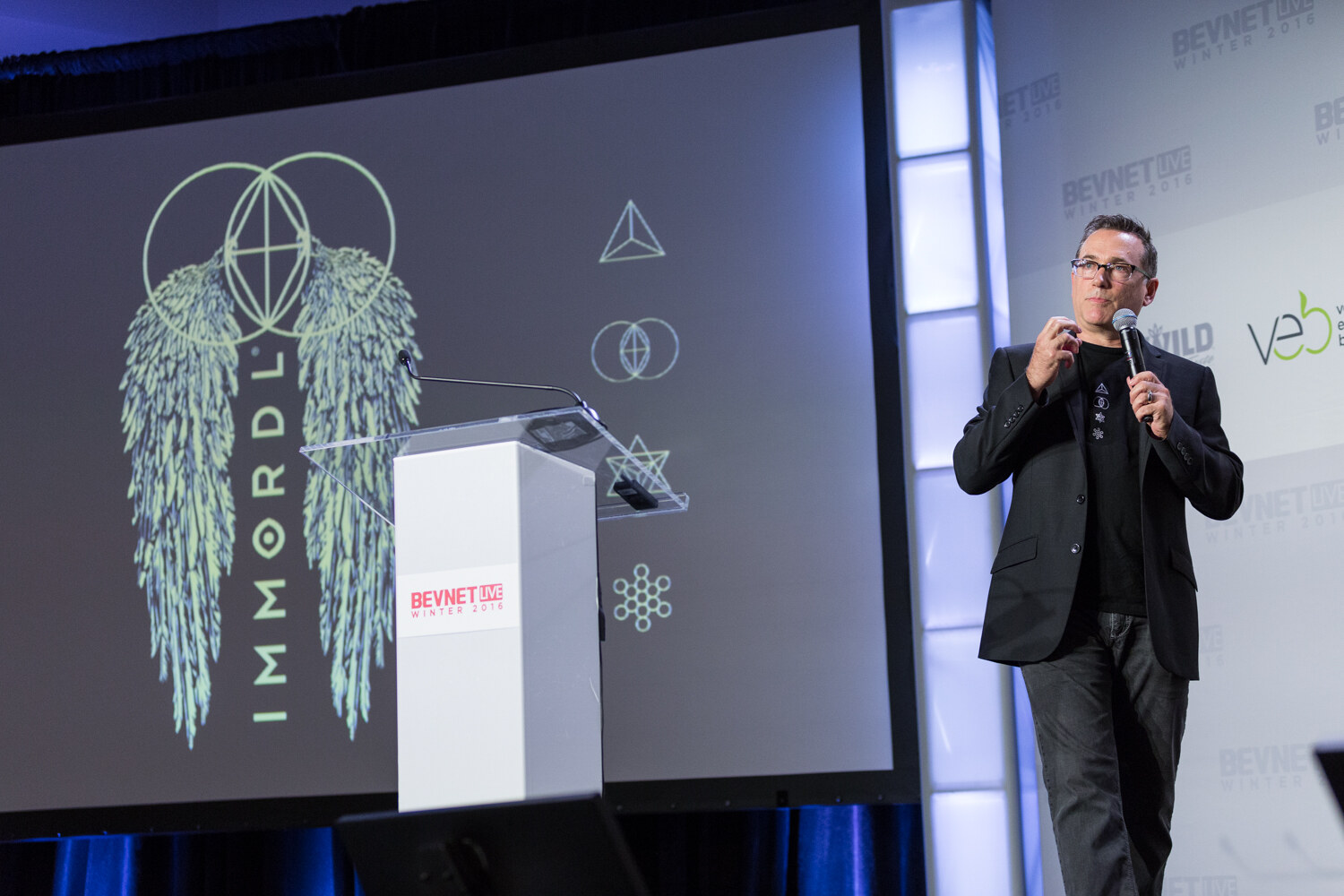 A man in glasses and a black blazer holds a microphone at stage right while the screen shows the IMMORDL logo with winged artwork and geometric symbols.