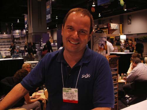 A smiling man in a navy polo shirt with a Blue Sky logo and a show badge stands in an exhibit hall, with other attendees seated at tables behind him.