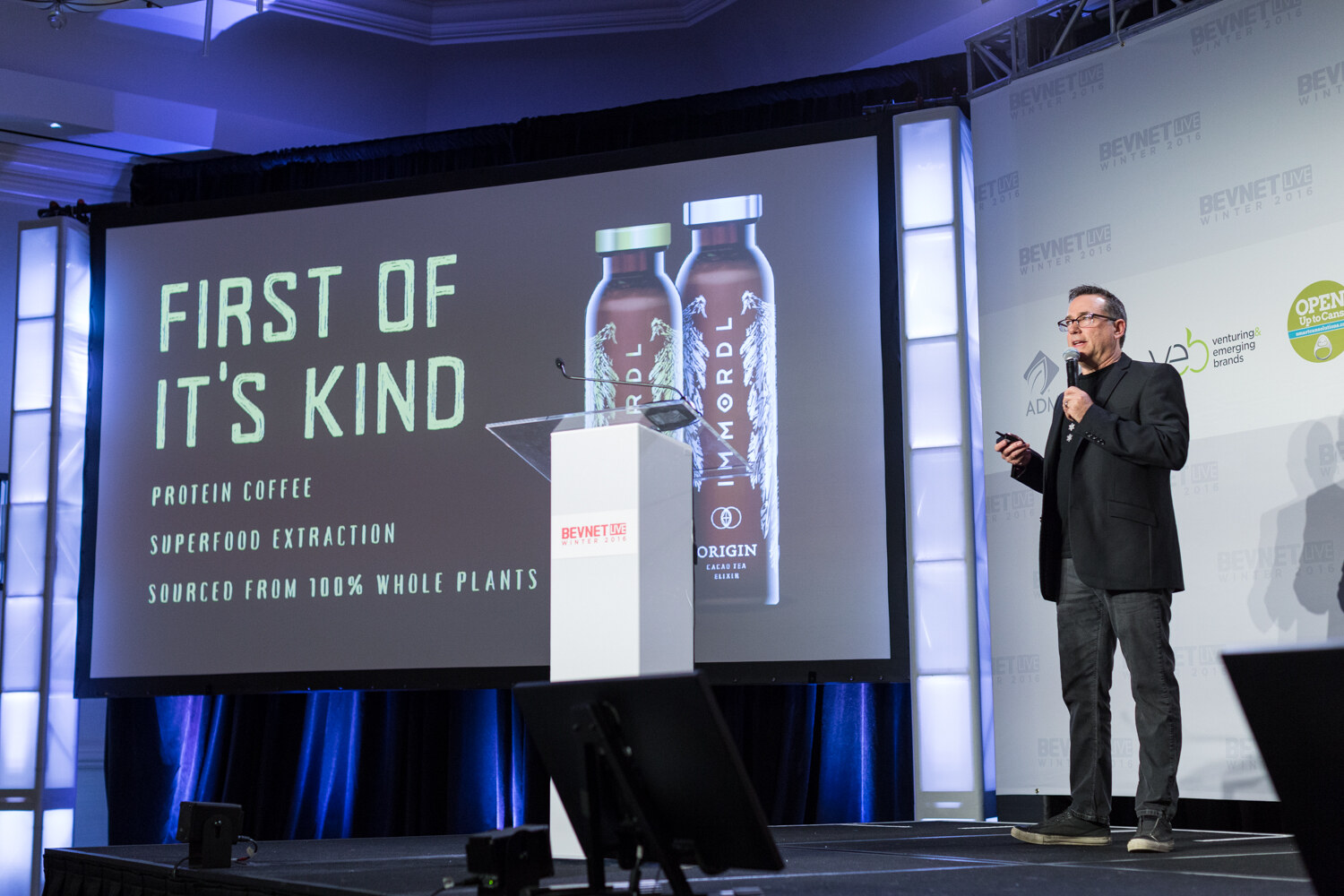 A man in glasses and a black blazer holds a microphone and clicker at stage right while the screen reads First of It's Kind: Protein Coffee, Superfood Extraction, Sourced from 100% Whole Plants beside two IMMORDL Origin bottles.