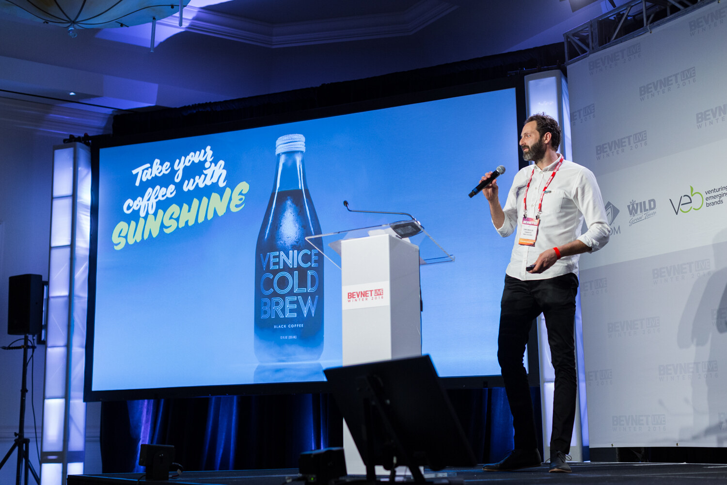 A bearded man in a white shirt and black pants holds a microphone on stage while the screen shows a Venice Cold Brew bottle and the line Take your coffee with sunshine.