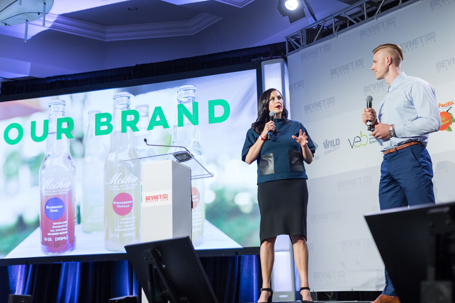 A woman in a teal top and black skirt and a man in a light blue shirt hold microphones on stage as the screen reads Our Brand above bottles labeled Mother, Blueberry Sage and Strawberry Vanilla.