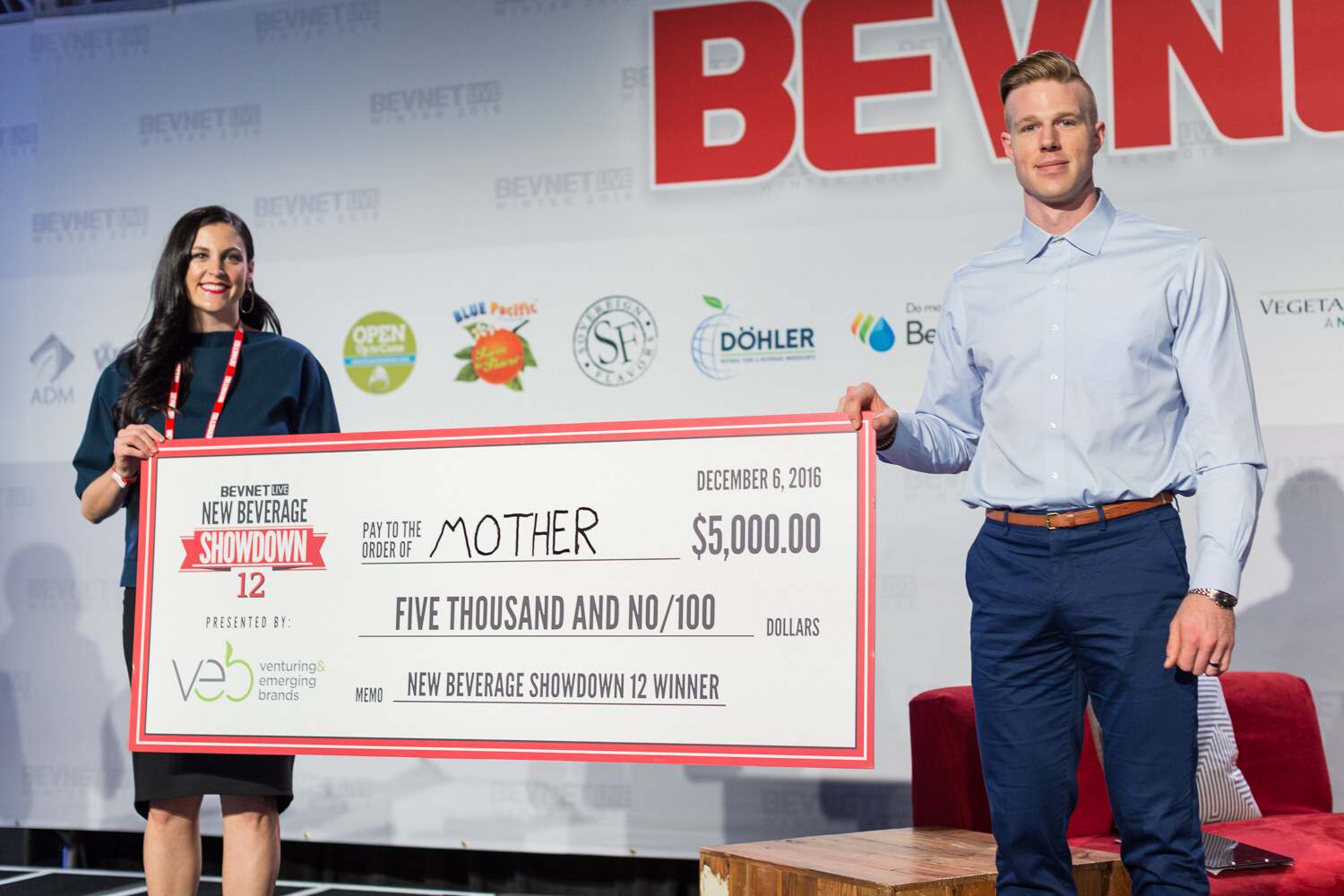 A woman in a teal top and a man in a light blue shirt hold a large check dated December 6, 2016 for $5,000 payable to Mother, marked New Beverage Showdown 12 Winner and presented by Venturing & Emerging Brands, in front of the sponsor wall.