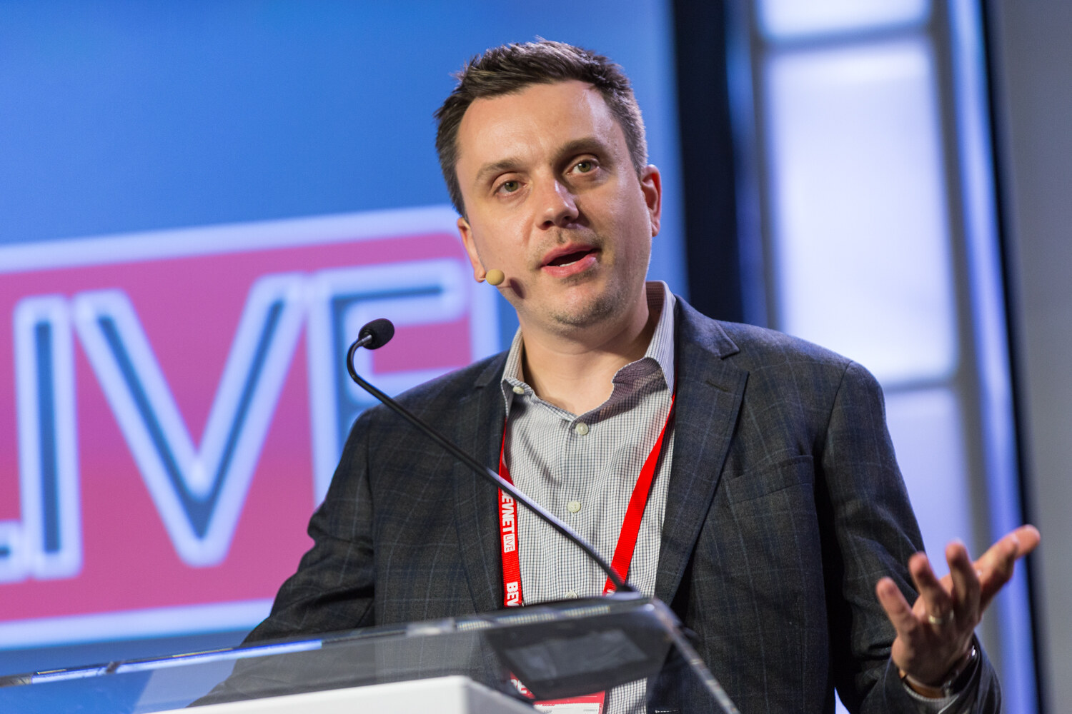 A man in a dark plaid blazer with a headset microphone and a red lanyard speaks at a clear lectern, one hand open, in front of a blue screen showing part of the BevNET Live logo.