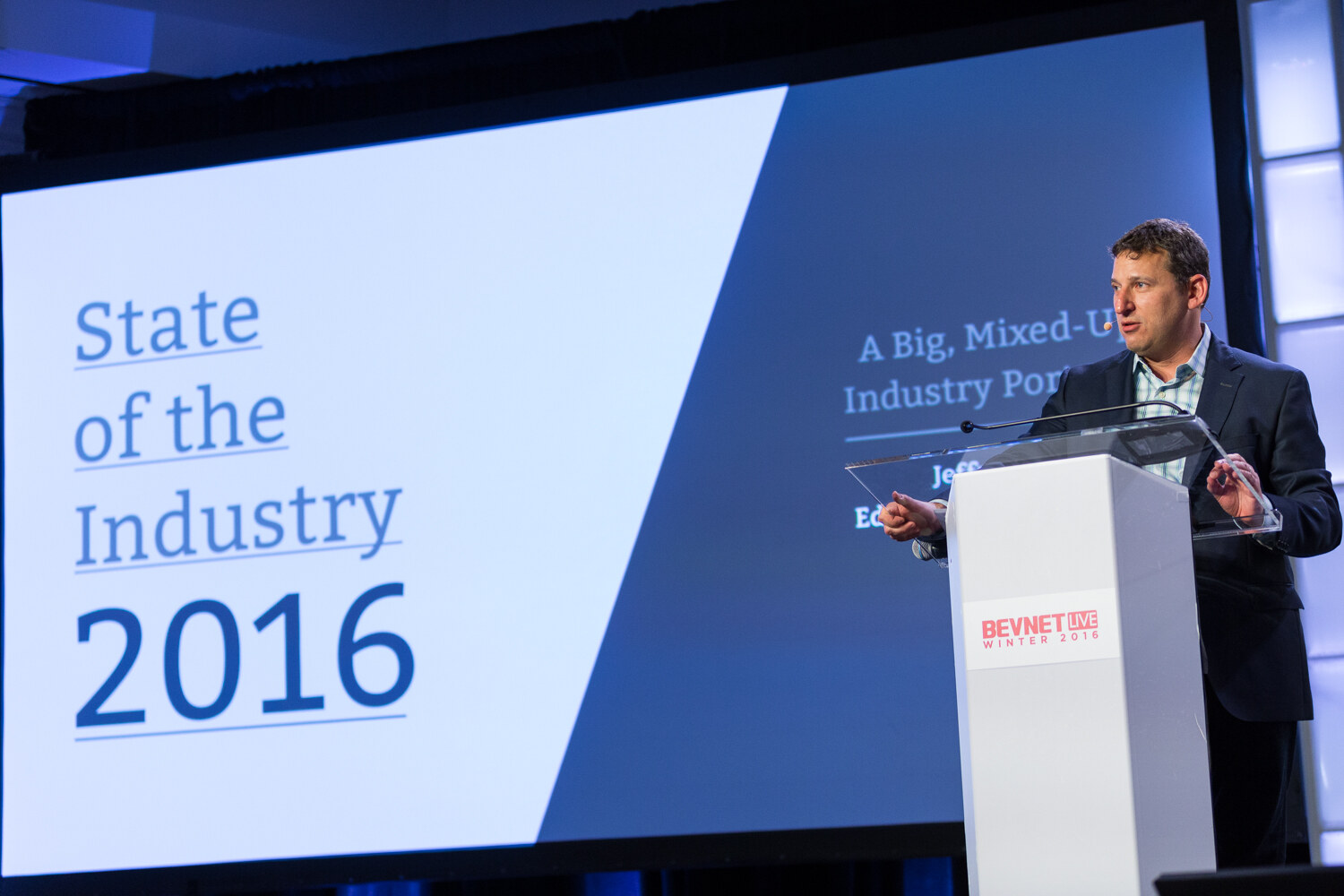 A man in a navy blazer speaks at a white BevNET Live Winter 2016 lectern next to a big screen reading State of the Industry 2016 and A Big, Mixed-Up Industry Portrait.