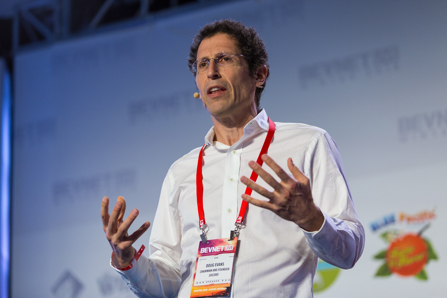 A man in glasses and a white shirt with a speaker badge on a red lanyard and a headset microphone holds both hands open in front of the BevNET Live backdrop.