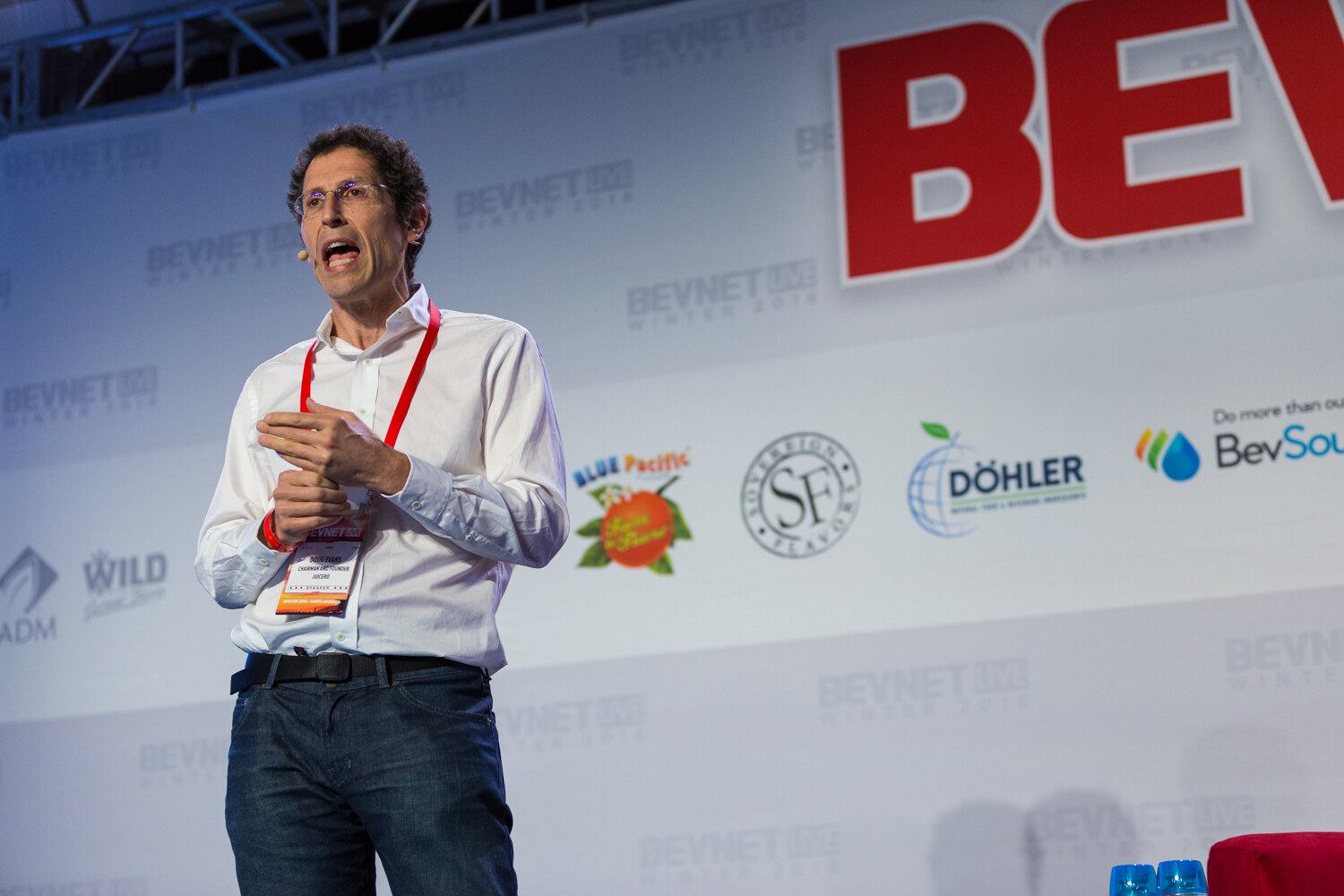 A man in a white shirt, jeans and speaker badge stands on stage with clasped hands in front of a backdrop with Blue Pacific, Sovereign Flavors, Dohler, BevSource, ADM and Wild logos.