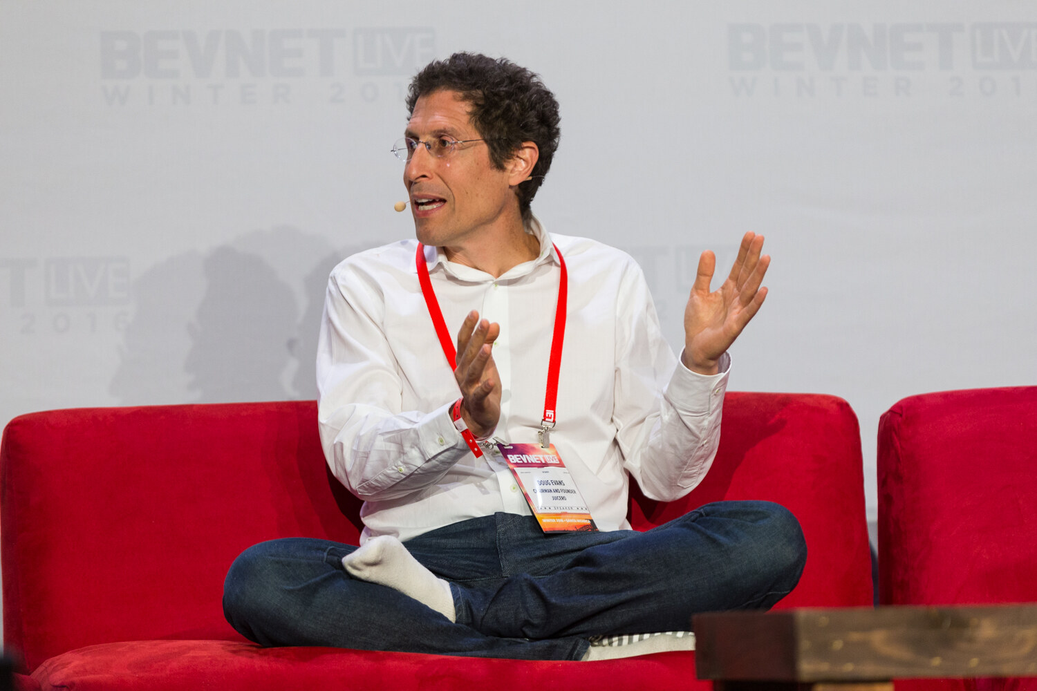 A man in a white shirt and jeans sits cross-legged on a red couch with both palms raised in front of a BevNET Live Winter 2016 backdrop.
