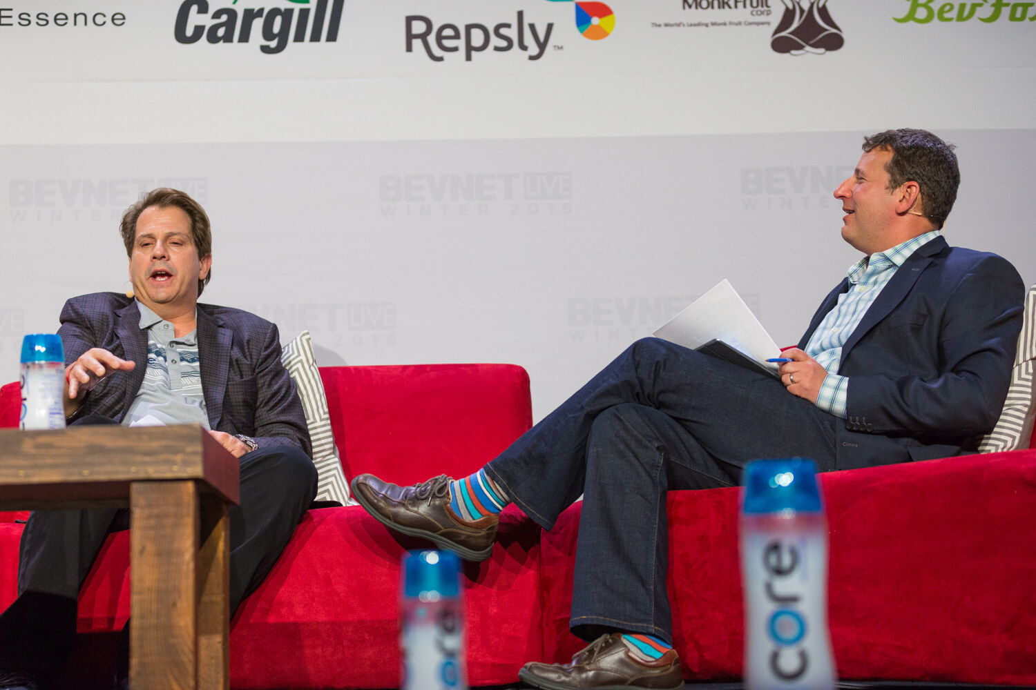 Two men sit on red couches under a sponsor wall with Cargill, Repsly and Monk Fruit logos; a Core water bottle stands on the table and on the stage floor.