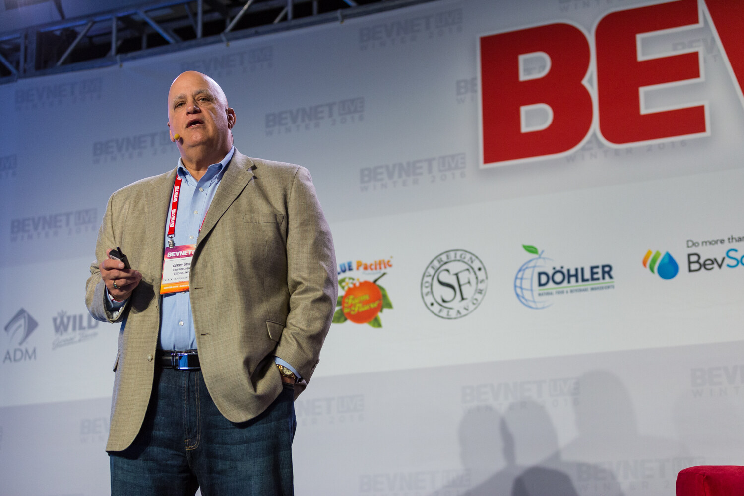 A bald man in a tan blazer and blue shirt with a speaker badge stands on stage with a clicker in his hand in front of a backdrop with ADM, Wild, Blue Pacific, Dohler and BevSource logos.