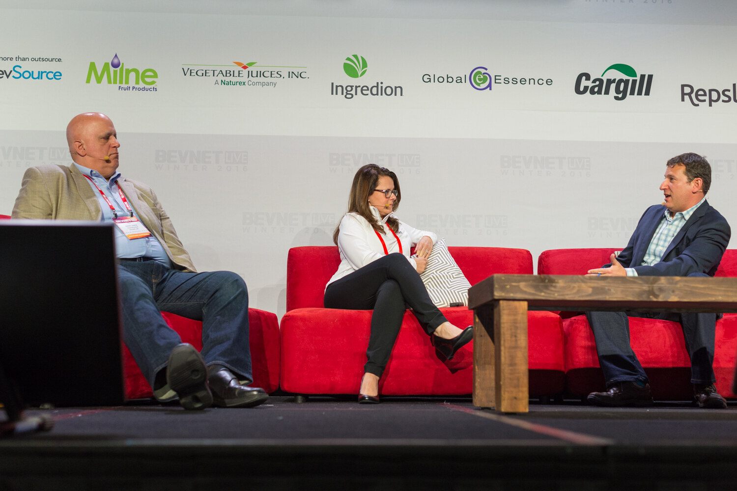 A bald man in a tan blazer, a woman in a white pullover and a man in a navy blazer sit on red couches under a backdrop with Milne, Vegetable Juices, Ingredion, Global Essence, Cargill and Repsly logos.