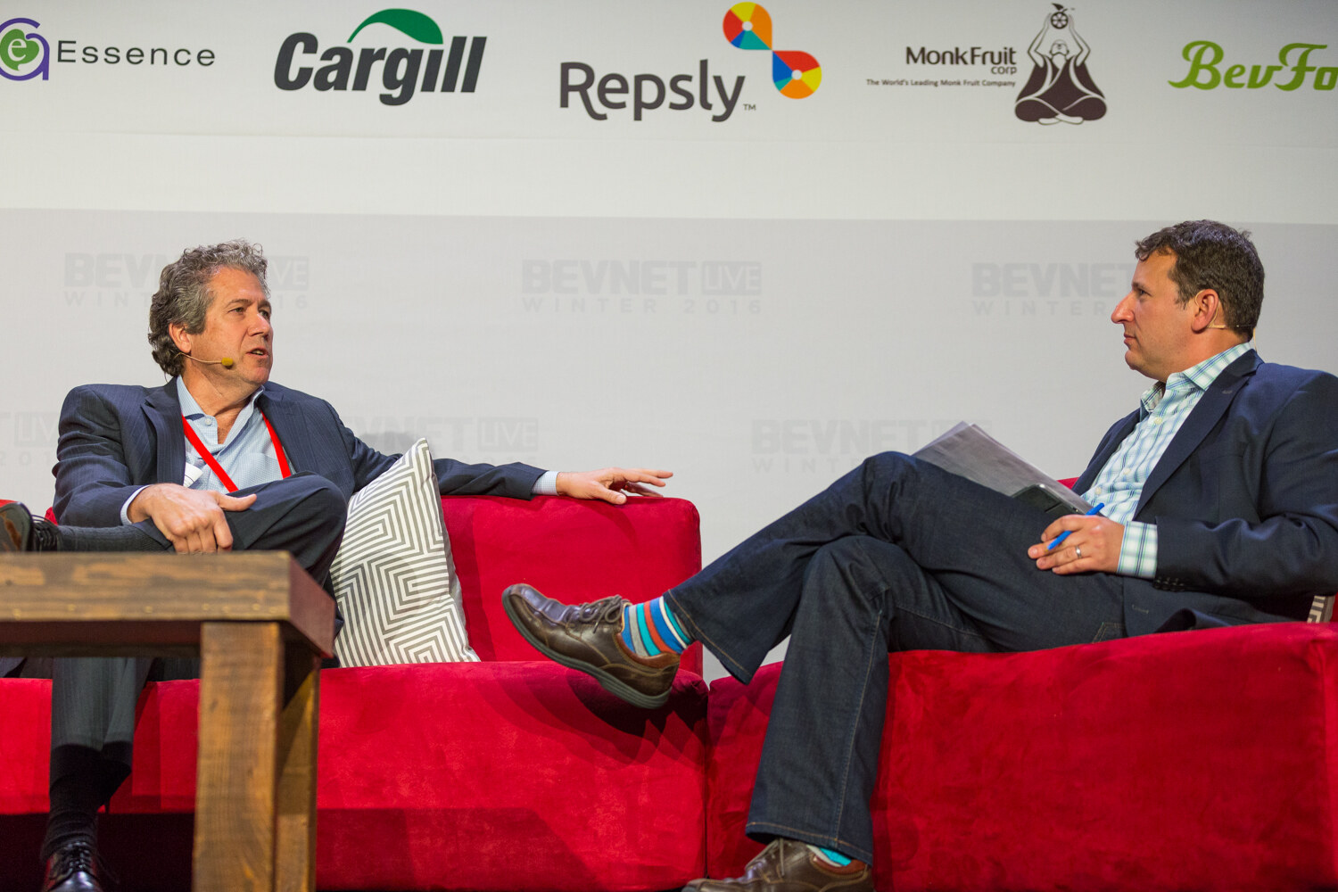 A man with curly gray hair in a navy blazer gestures with one hand while seated opposite a moderator in a blue checked shirt holding notes, under a sponsor wall with Cargill, Repsly and Monk Fruit logos.