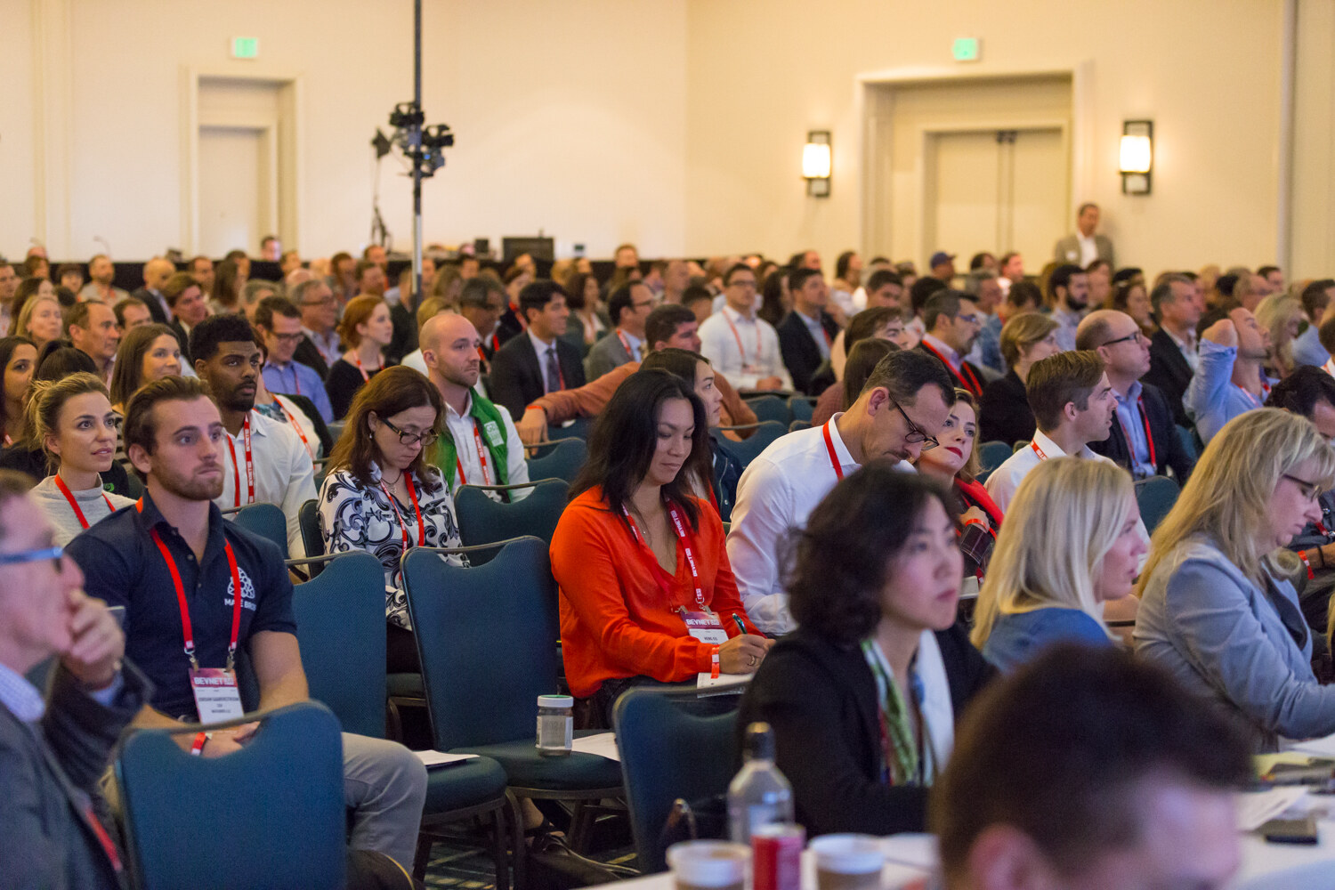 Rows of seated attendees with red BevNET Live lanyards face the stage in a hotel ballroom; a woman in an orange top takes notes in the foreground.