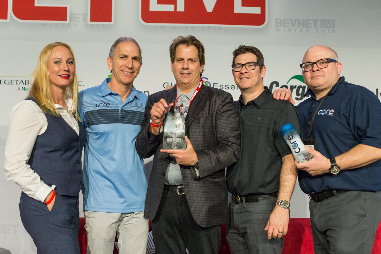 Five people stand in a row; two wear Core logo polos, the man in the middle holds a glass trophy and another holds a Core water bottle, in front of the sponsor wall.