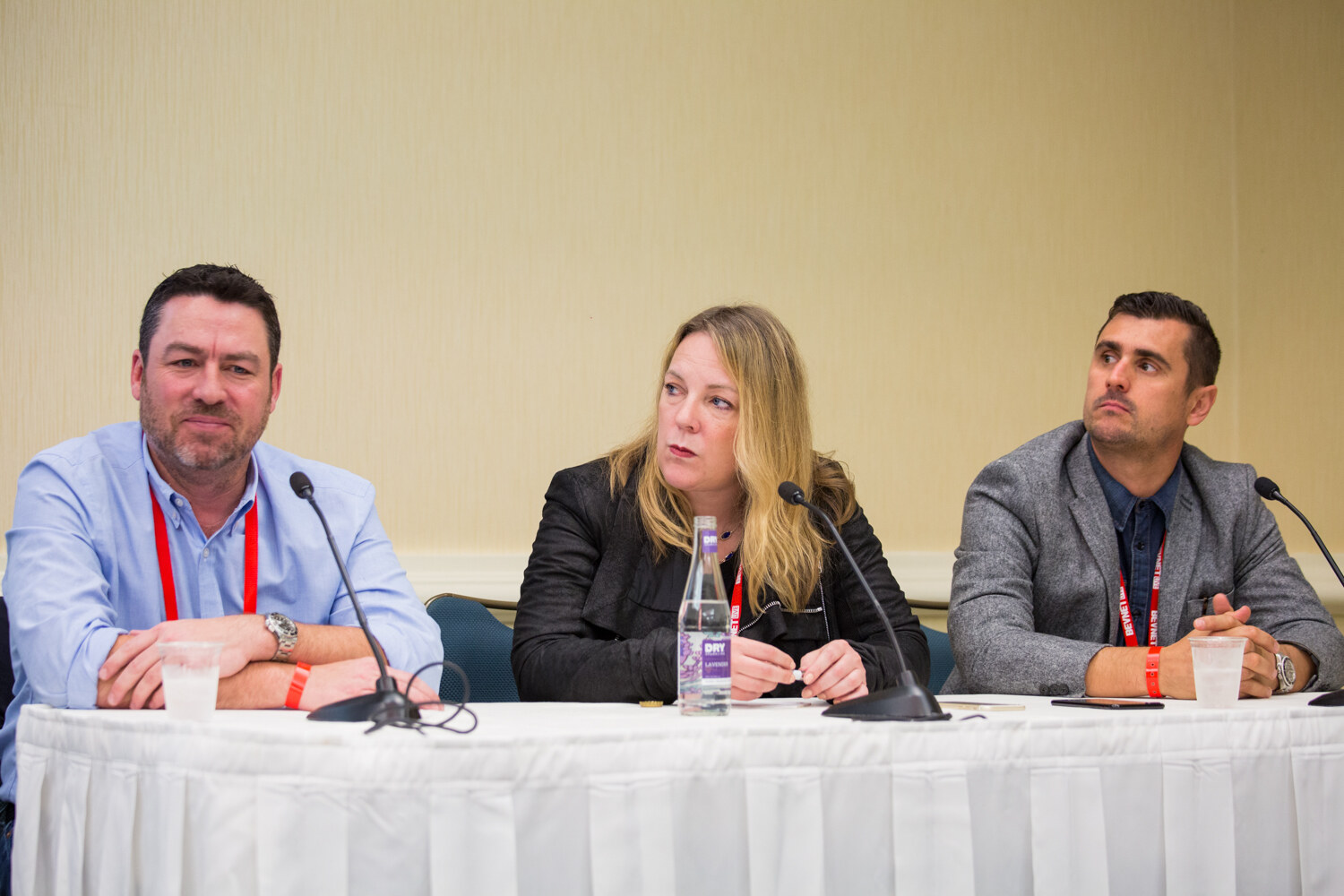 Two men and a woman with red lanyards sit behind a white skirted table with microphones, plastic cups and a glass water bottle in a hotel meeting room.