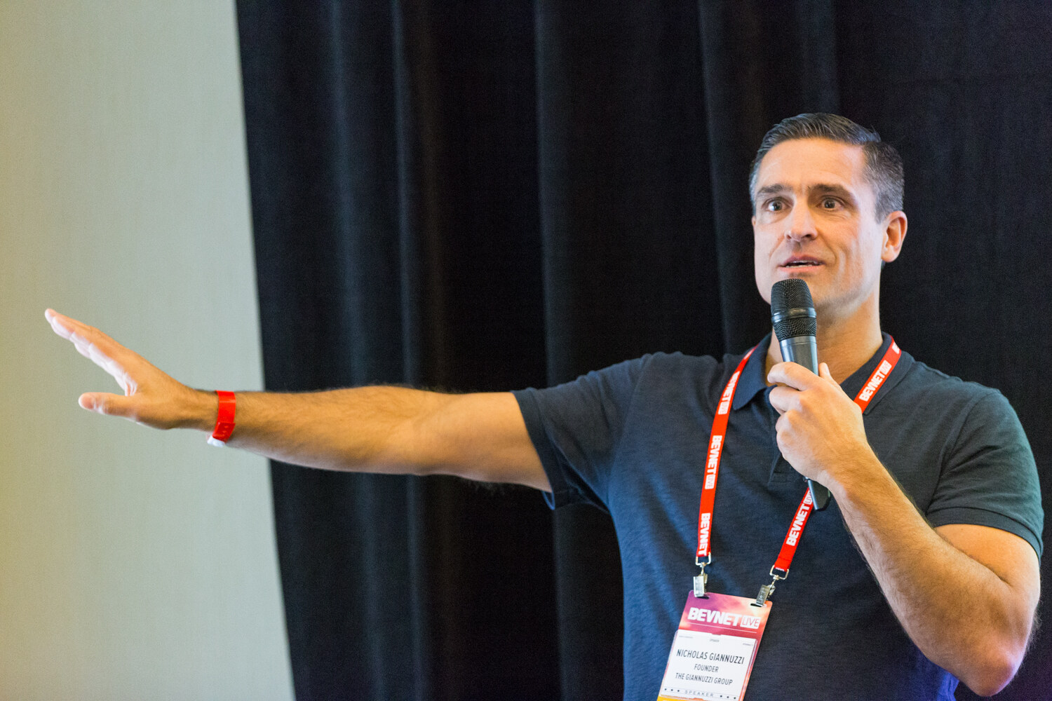 A man in a navy polo with a red lanyard and a speaker badge holds a handheld microphone and stretches his left arm out in front of a black curtain.