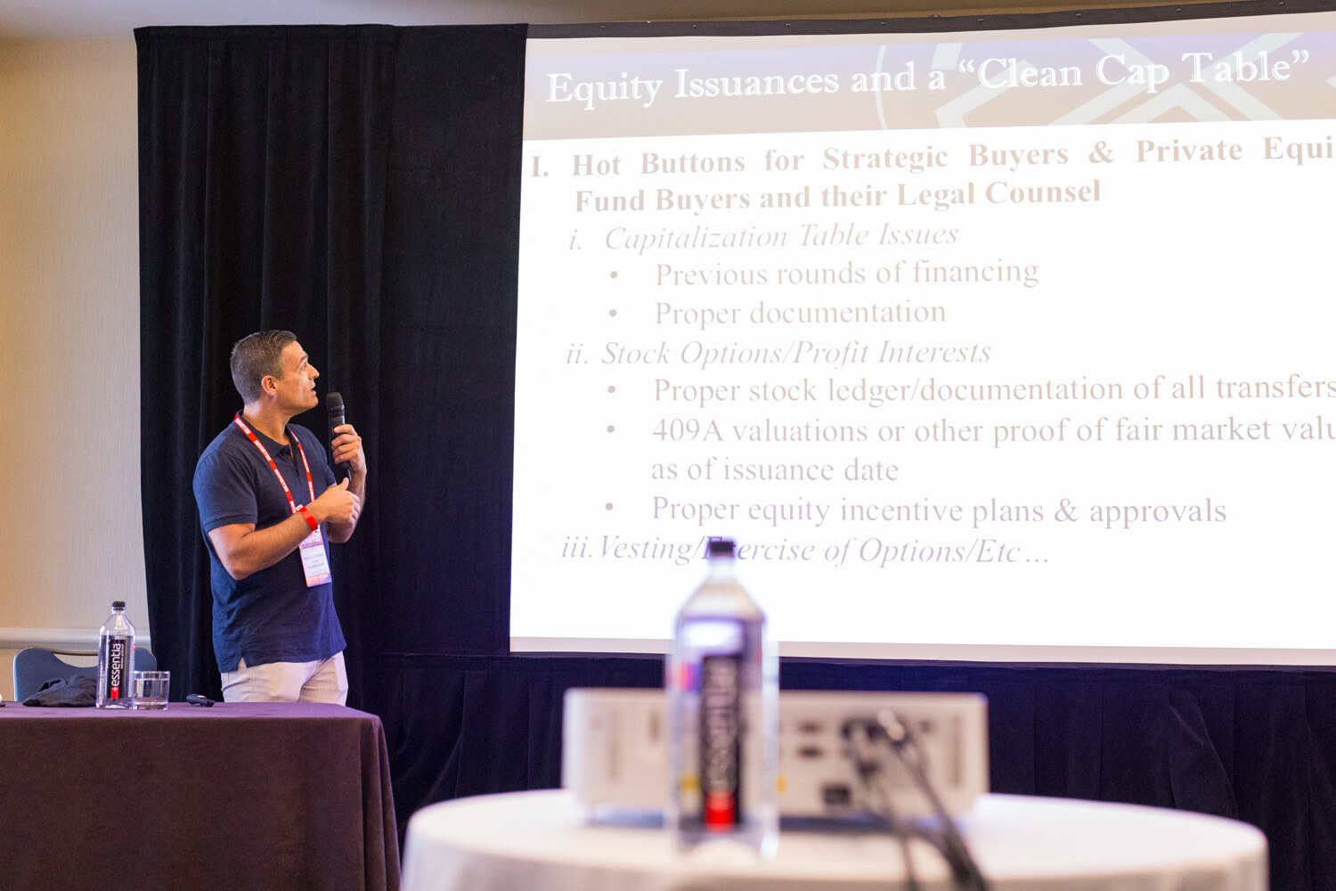 A man in a navy polo holding a microphone looks at a projected slide titled Equity Issuances and a Clean Cap Table, with Essentia water bottles on the tables.