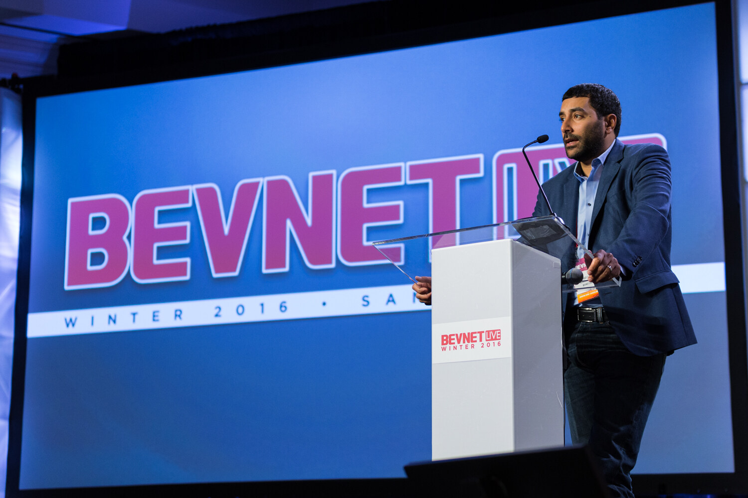 A man in a navy blazer stands at a white lectern marked BevNET Live Winter 2016 in front of a large blue screen showing the BevNET Live Winter 2016 Santa Monica logo.