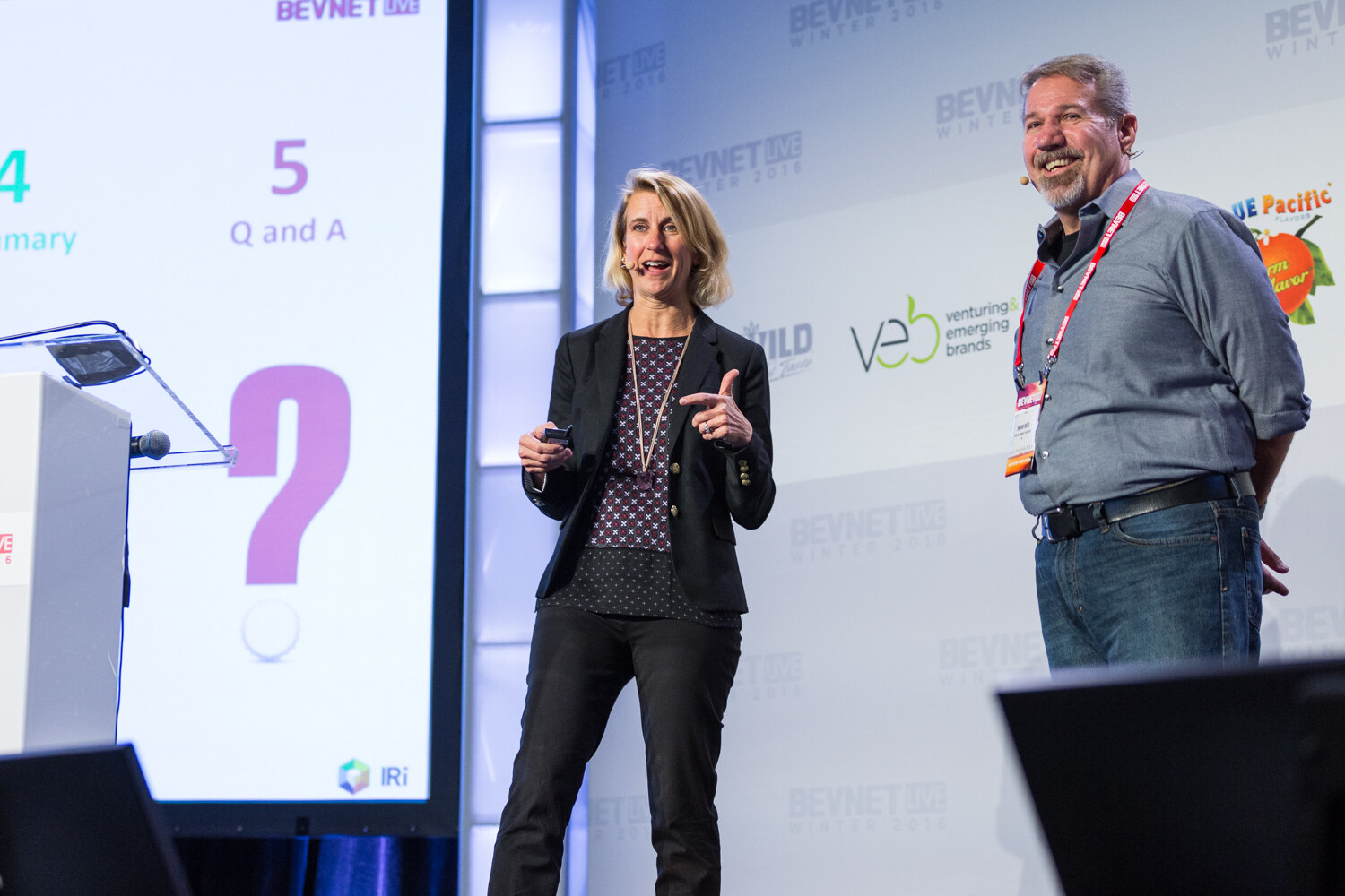 A woman in a black blazer holding a clicker points forward while a man in a gray shirt stands beside her; the screen shows a question mark, Q and A and an IRI logo.