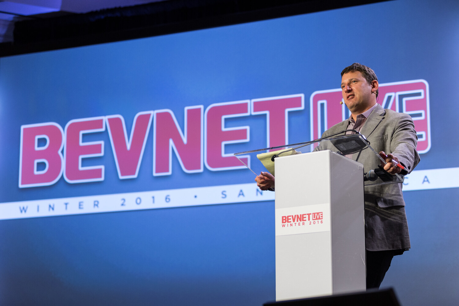 A man in a gray blazer with a headset microphone holds a yellow legal pad and a red pen at a white lectern marked BevNET Live Winter 2016, in front of a blue screen reading BevNET Live Winter 2016 Santa Monica.