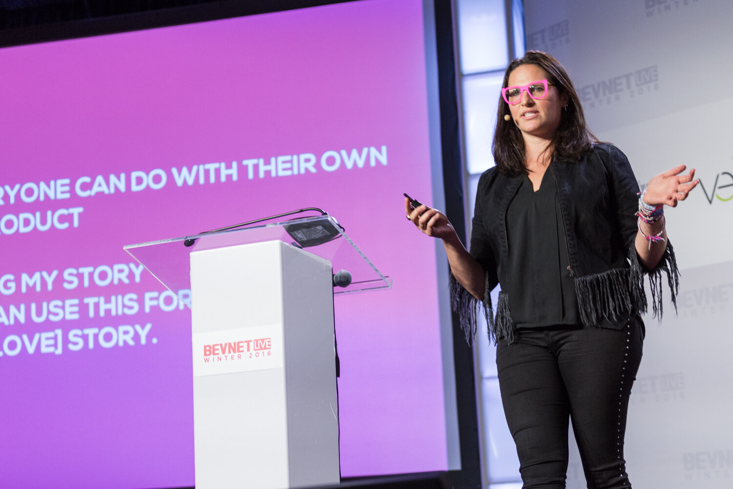 A woman in pink-framed glasses, a black fringed jacket and black pants holds a clicker and gestures beside a white lectern, in front of a magenta slide with the words Everyone can do with their own product and My story.