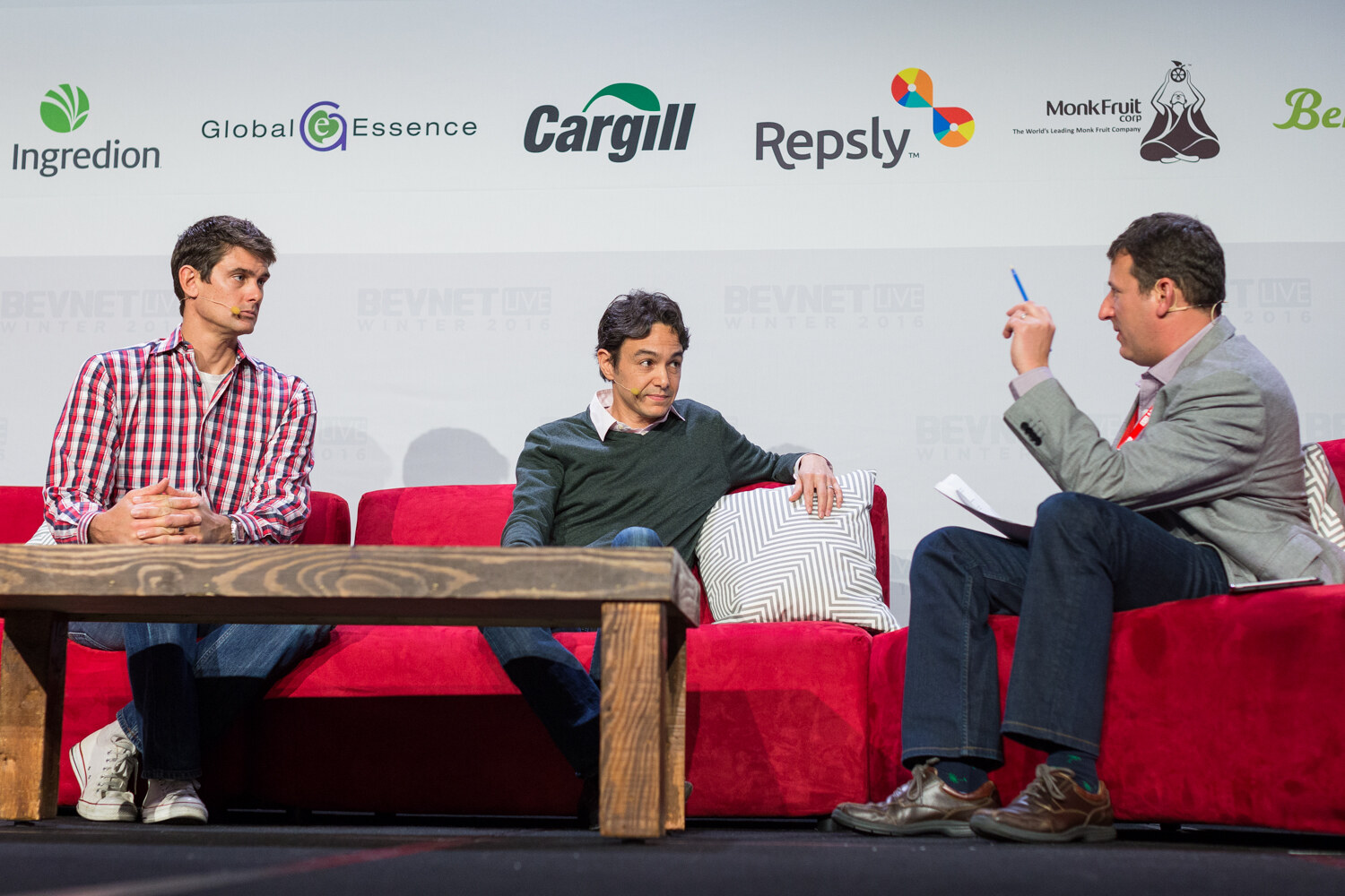 Two men on a red couch, one in a red plaid shirt and one in a green sweater, face a moderator in a gray blazer who points with a pen, under a wall of sponsor logos including Ingredion, Global Essence and Cargill.