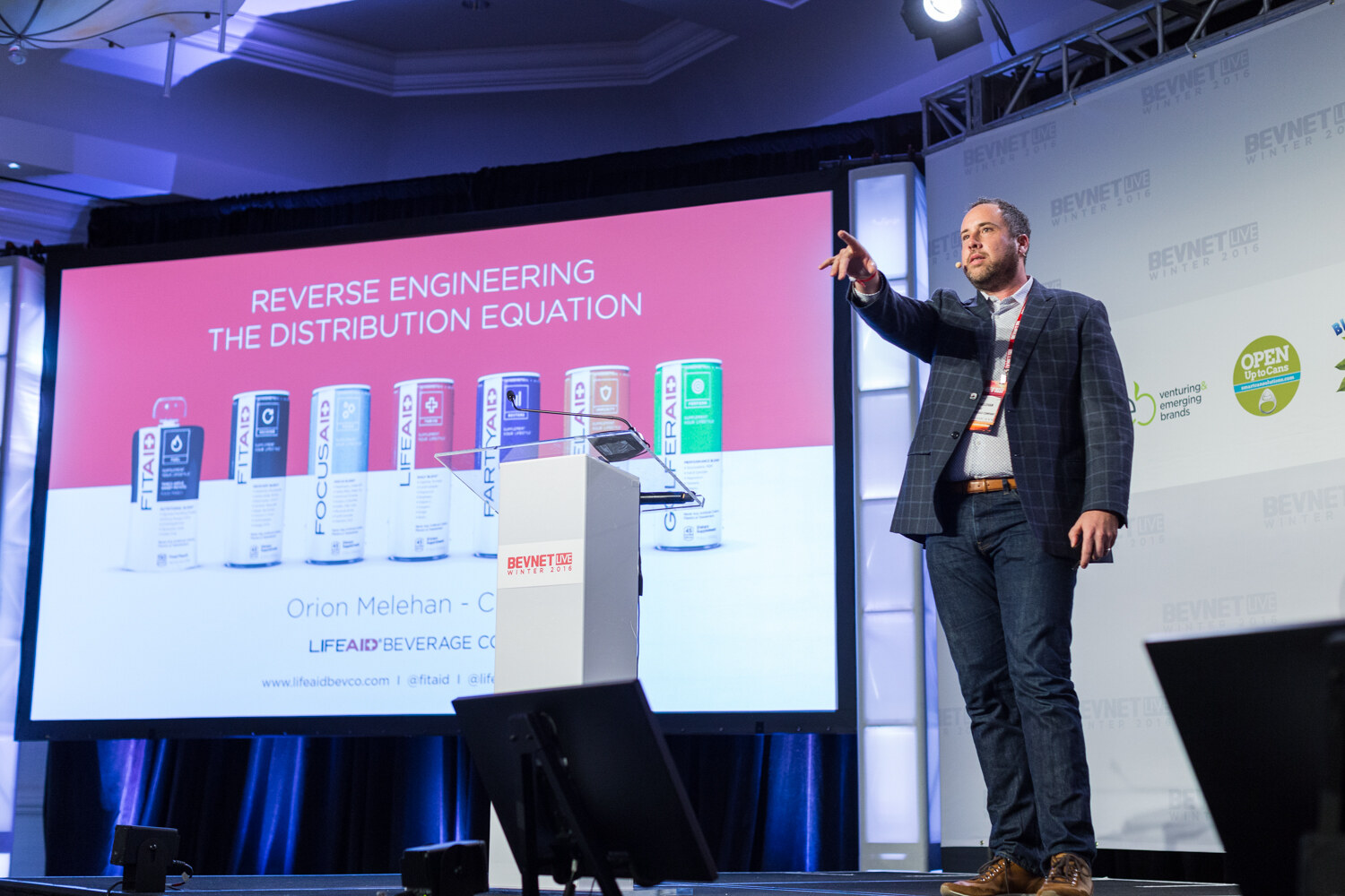 A man in a navy plaid blazer and jeans stands at stage right with his arm extended; the screen reads Reverse Engineering the Distribution Equation above cans labeled FITAID, FOCUSAID, PARTYAID and LIFEAID.