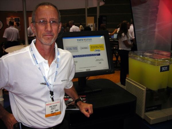 Dave Tuchler of Tate & Lyle, wearing glasses, a white polo and a Tate & Lyle lanyard, stands by a monitor showing the Tate & Lyle logo, next to a dispenser of yellow-green drink.