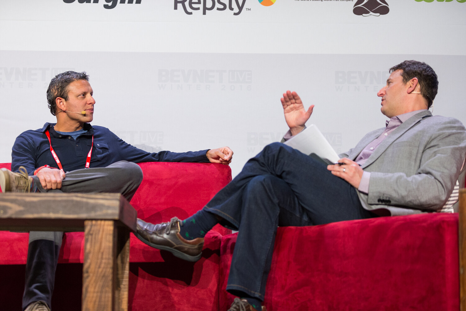 A man in a navy pullover with a red lanyard and headset microphone rests his arm on a red couch while a moderator in a gray blazer gestures in conversation, with Repsly and Monk Fruit logos above.