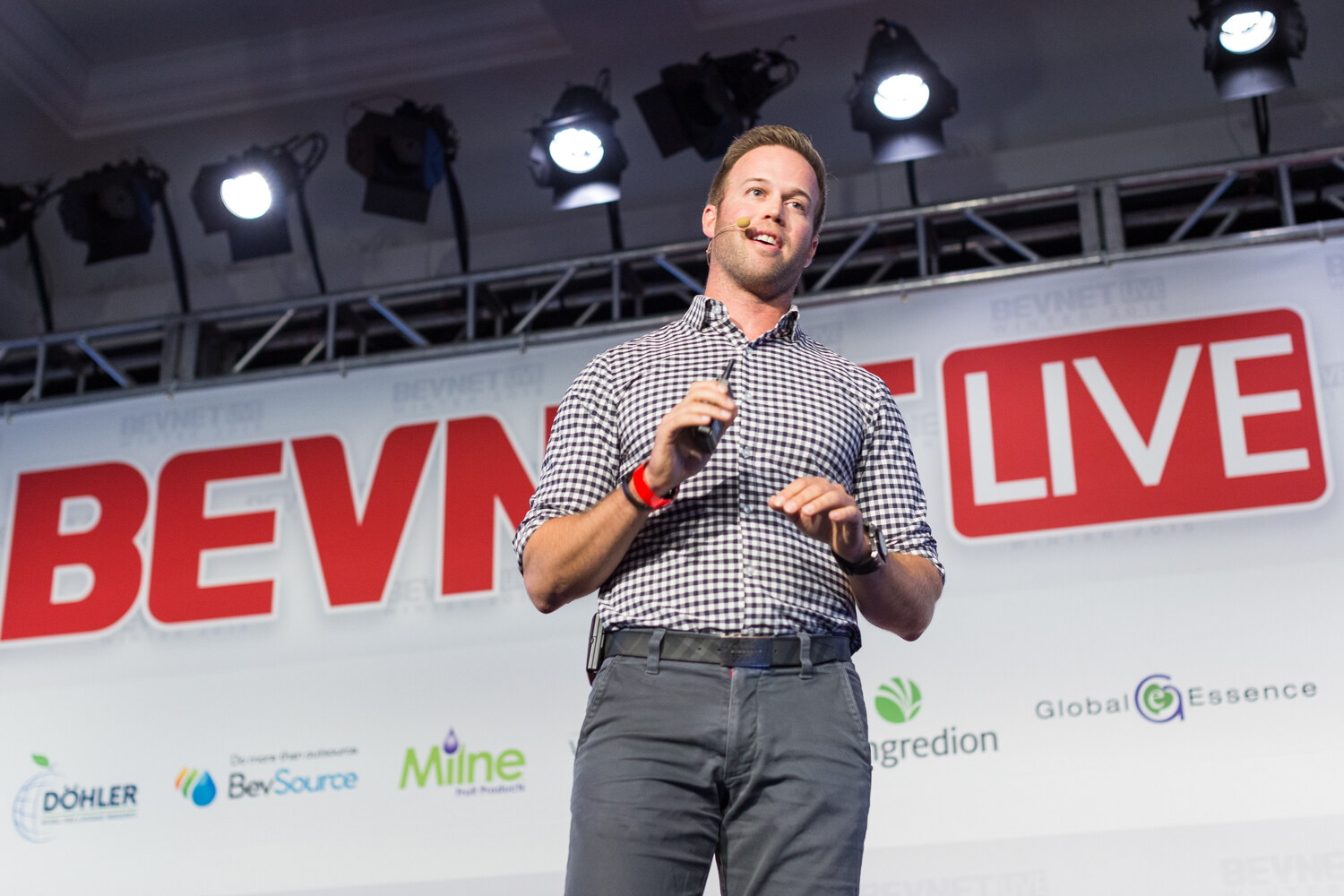A man with a headset microphone in a black and white gingham shirt and gray pants stands on stage in front of a large red BevNET Live sign with Dohler, BevSource, Milne, Ingredion and Global Essence logos below.