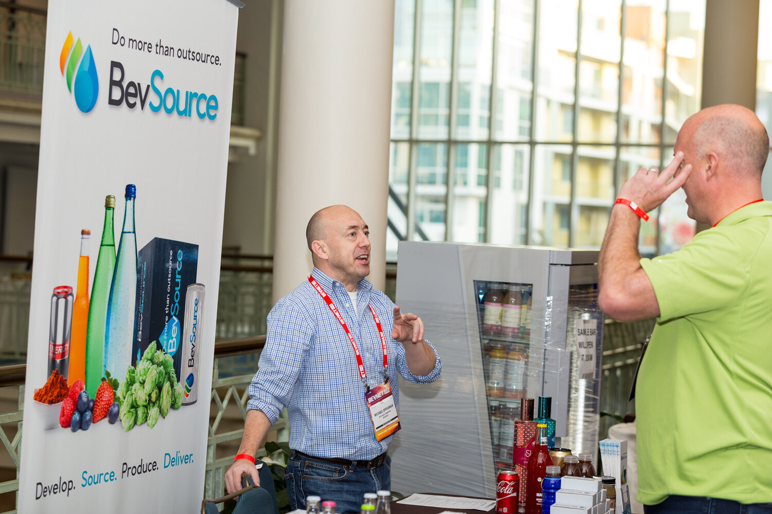 A bald man in a blue checked shirt gestures while talking to a visitor in a green polo, beside a BevSource banner reading Do more than outsource and a small glass-door fridge of beverages.