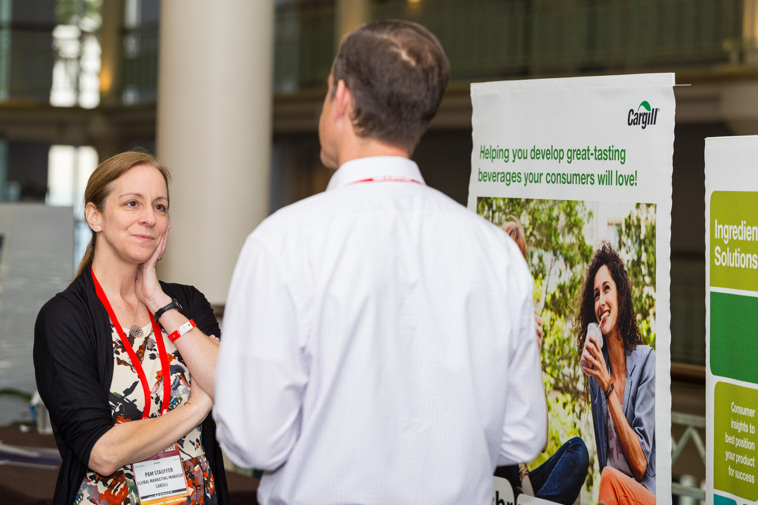 A woman with a red BevNET Live lanyard rests her chin on her hand while talking with a man seen from behind, beside a Cargill banner reading Helping you develop great-tasting beverages your consumers will love.