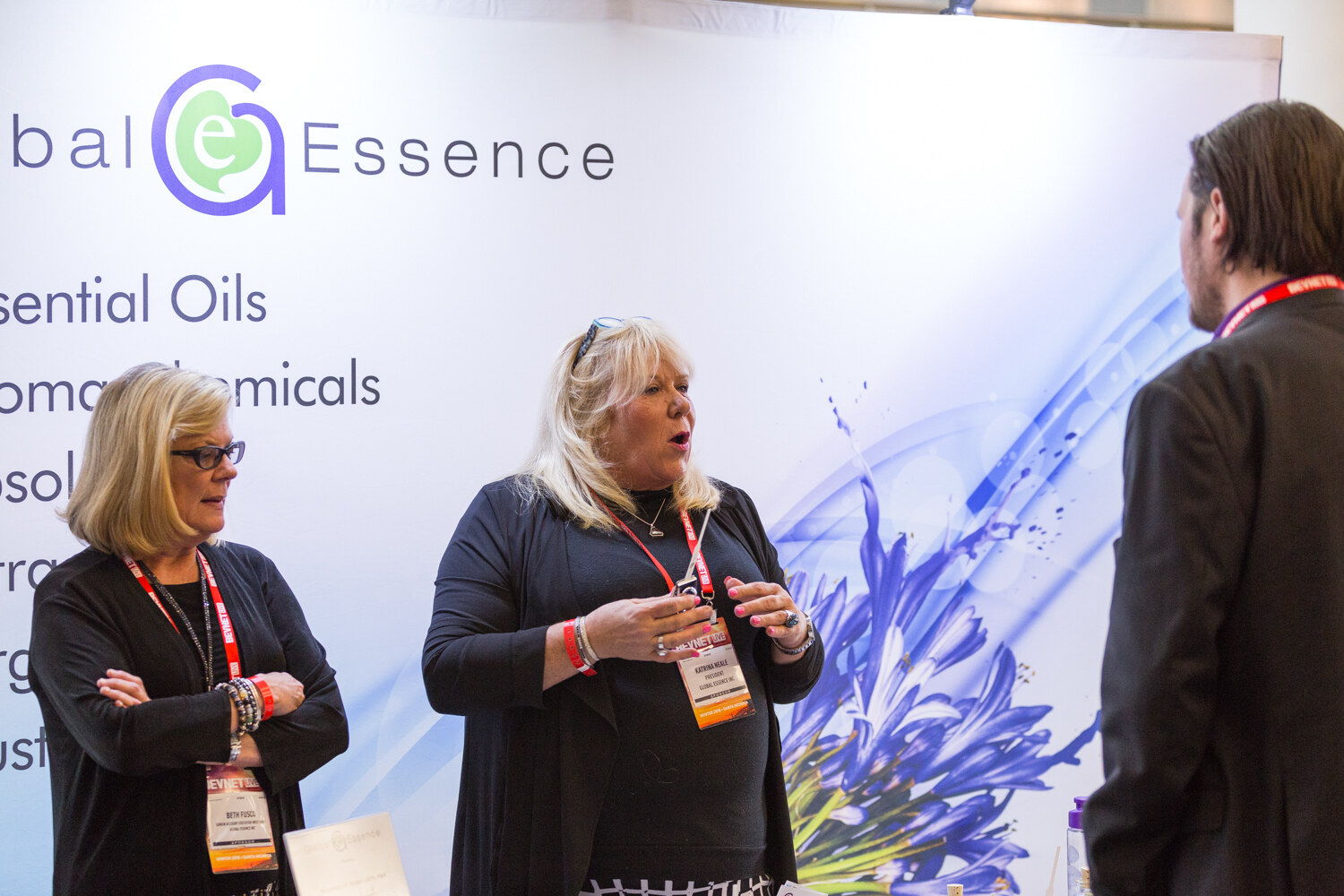 Two women in black with BevNET Live badges stand in front of a Global Essence backdrop listing essential oils and aroma chemicals while one speaks to a man seen from behind.