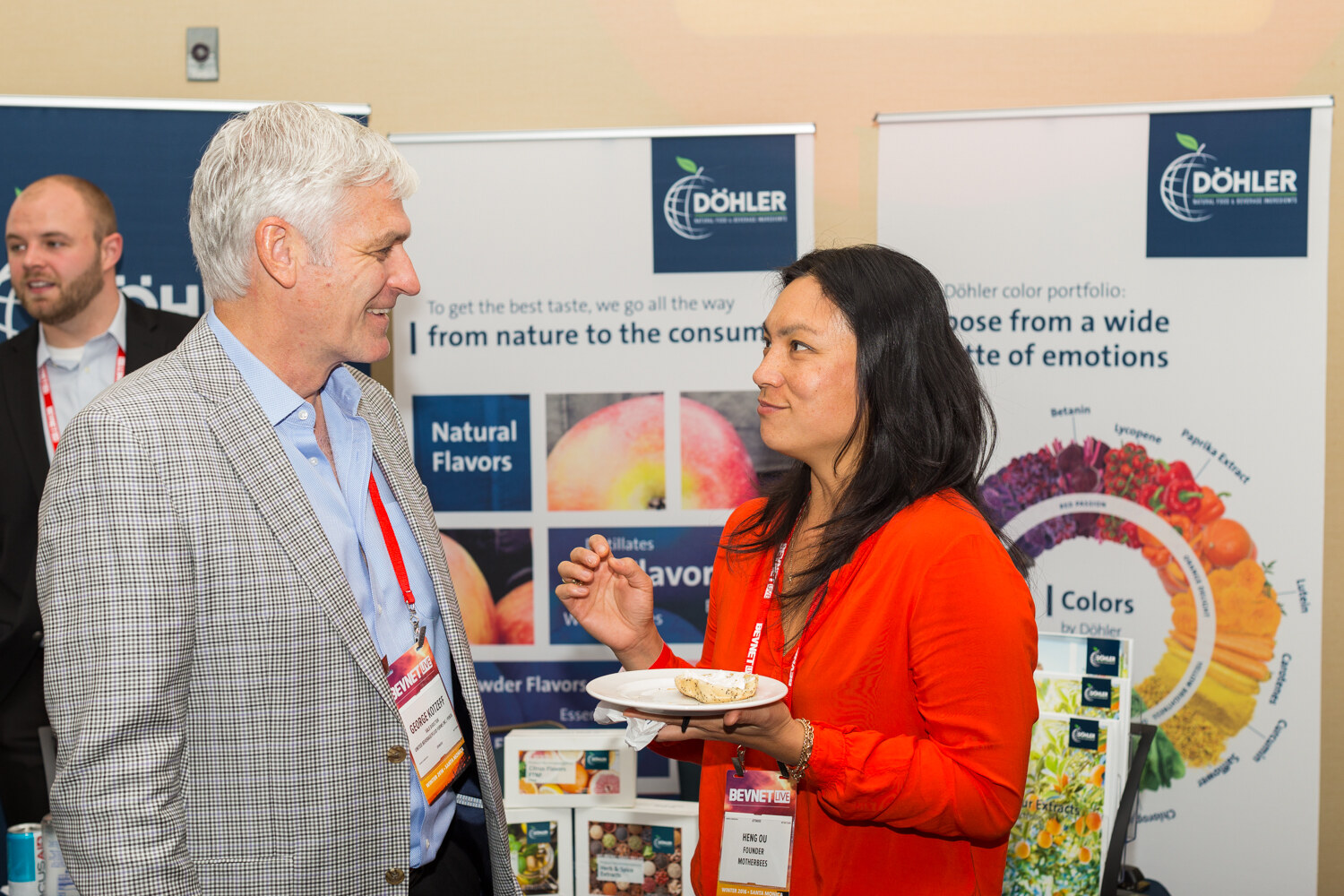 A gray-haired man in a houndstooth blazer talks with a woman in an orange blouse holding a plate, with Dohler banners about natural flavors and colors behind them.
