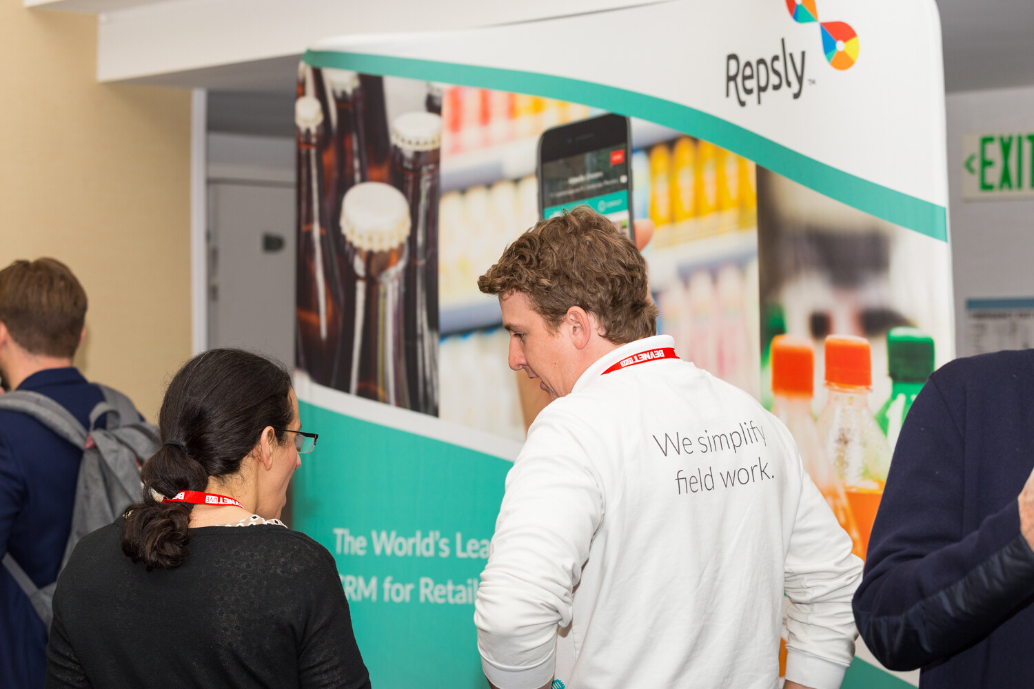 A man in a white sweatshirt printed We simplify field work stands in front of a green and white Repsly display with bottle photos, speaking with a woman with glasses seen from behind.