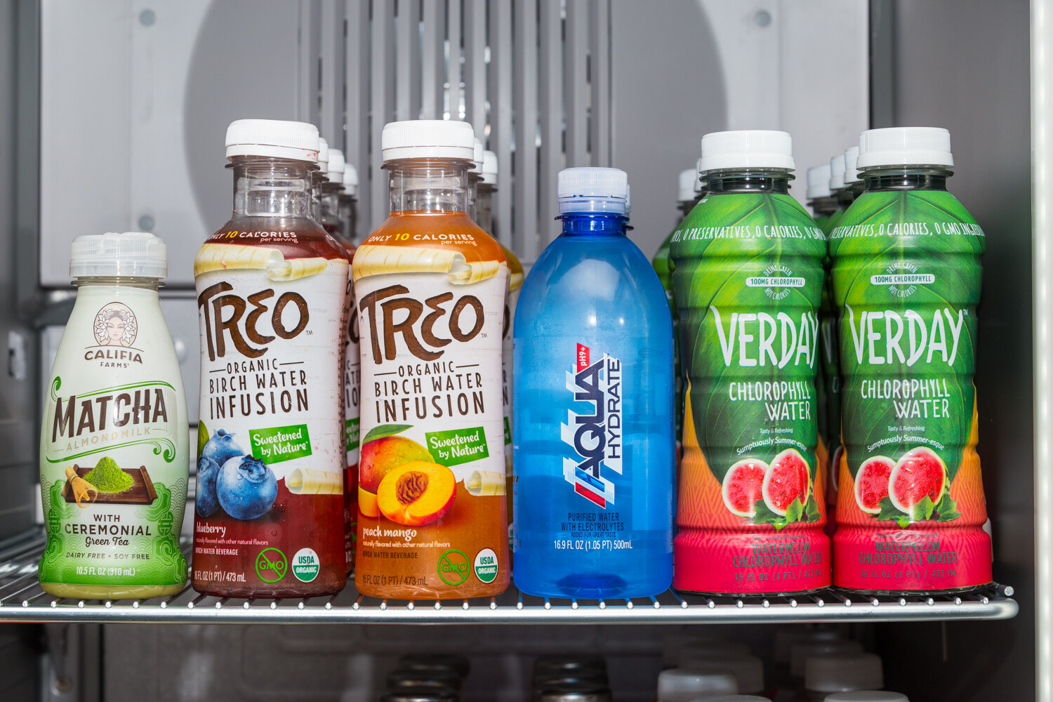 Close view of a refrigerator shelf with Califia Farms Matcha almondmilk, two Treo Organic Birch Water Infusion bottles, an Aqua Hydrate bottle and two Verday Chlorophyll Water bottles.