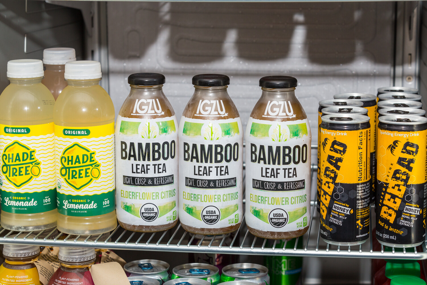 Two Shade Tree Original Lemonade bottles, three Igzu Bamboo Leaf Tea Elderflower Citrus bottles and a stack of yellow and black Beebad sparkling energy drink cans on a metal fridge shelf.