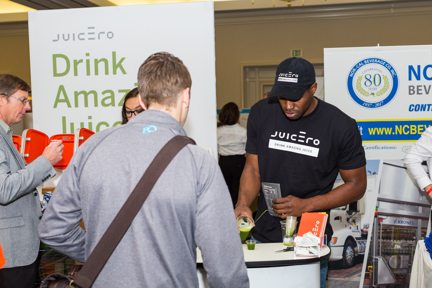 A man in a black Juicero cap and T-shirt pours green juice into a small cup at a white counter while a visitor with a shoulder bag looks on, with a Juicero Drink Amazing Juice banner behind them.