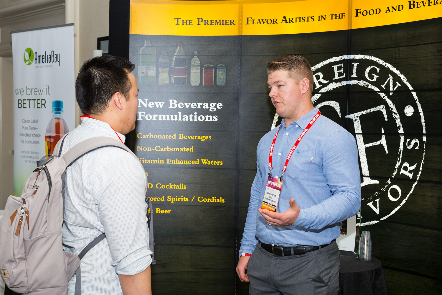 A man in a light blue shirt and red lanyard speaks with a man in a gray backpack, in front of a black Sovereign Flavors display listing new beverage formulations.