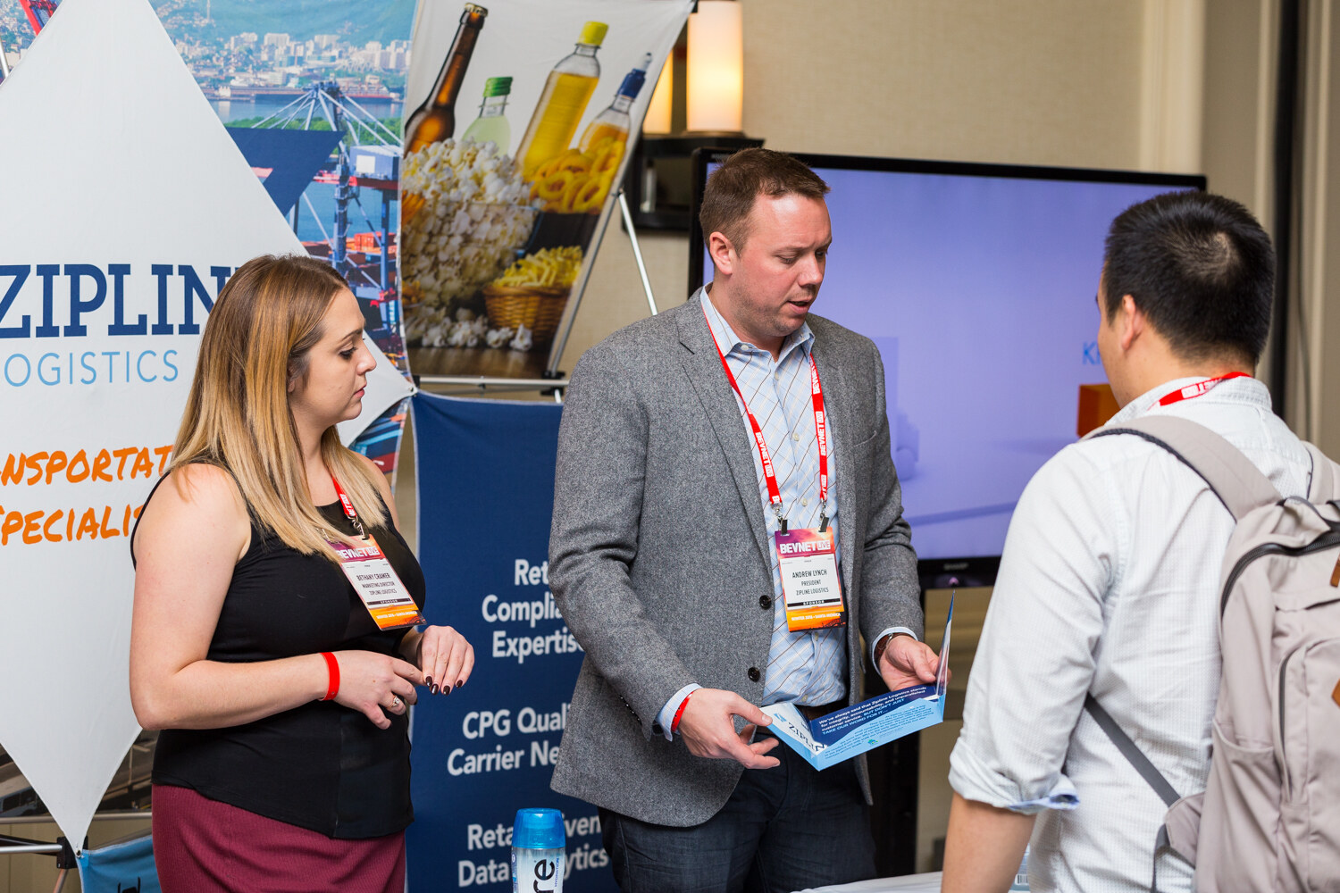 A man in a gray blazer holds a blue brochure while a woman in a black top looks on and a visitor in a backpack watches, with a Zipline Logistics banner about CPG carrier network behind them.