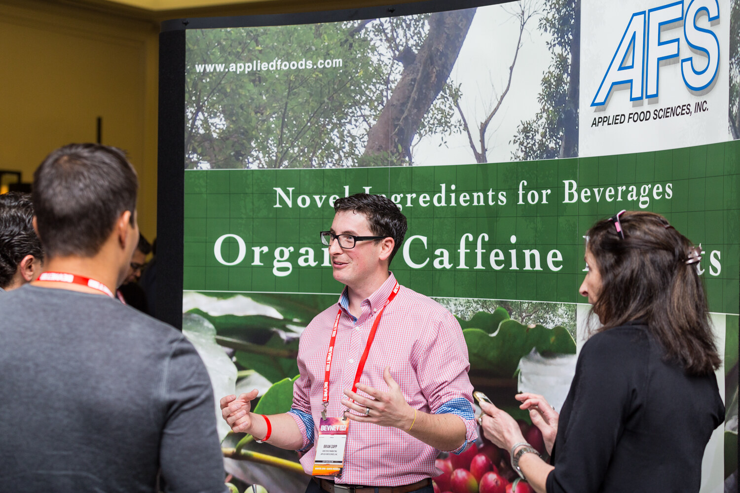A man in glasses and a pink gingham shirt with a red lanyard gestures while talking with a man seen from behind and a woman holding a phone; the backdrop reads Applied Food Sciences, Novel Ingredients for Beverages, Organic Caffeine.