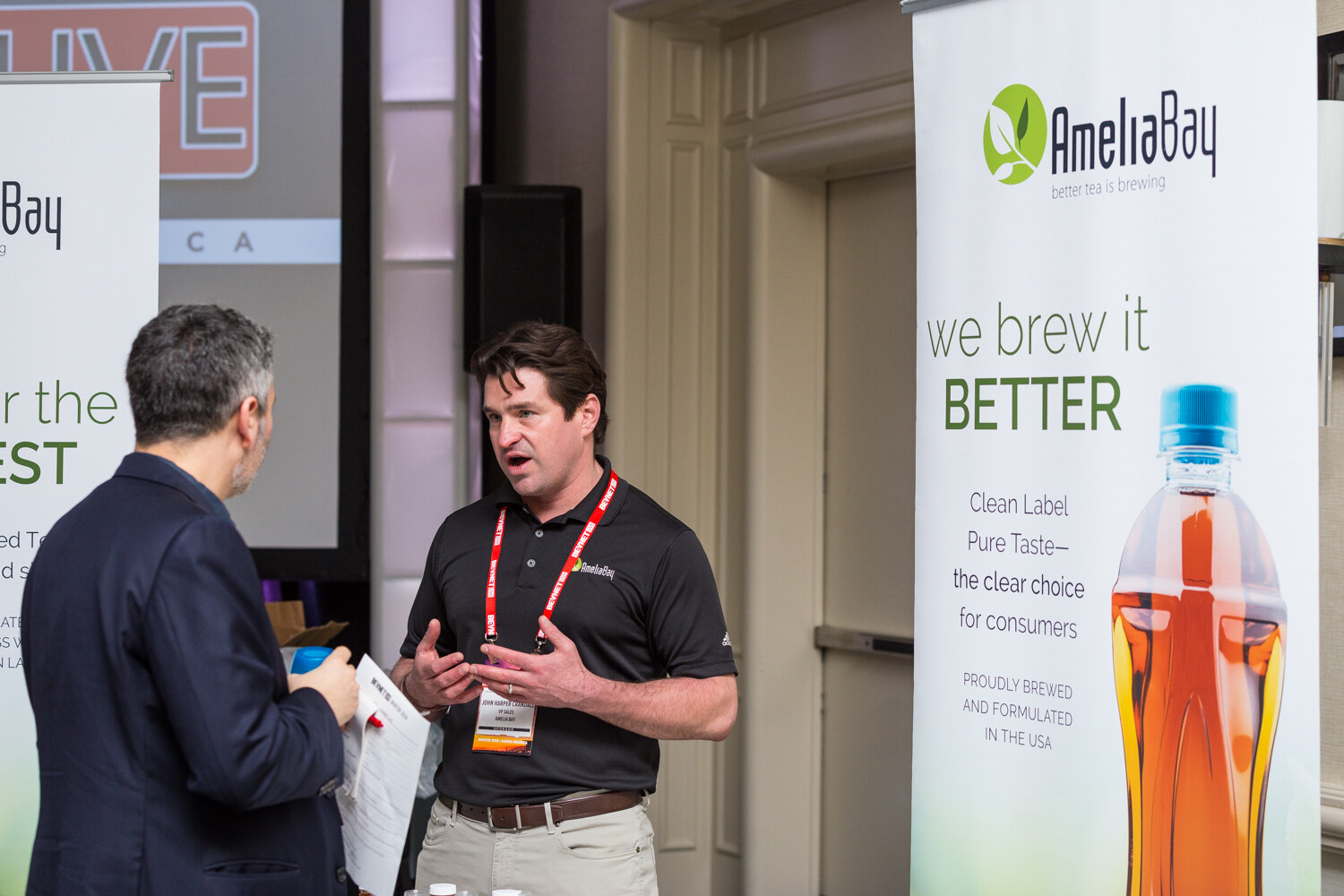 A man in a black AmeliaBay polo with a red lanyard speaks with a man holding a notepad, beside an AmeliaBay banner with an iced tea bottle and the line We brew it better.
